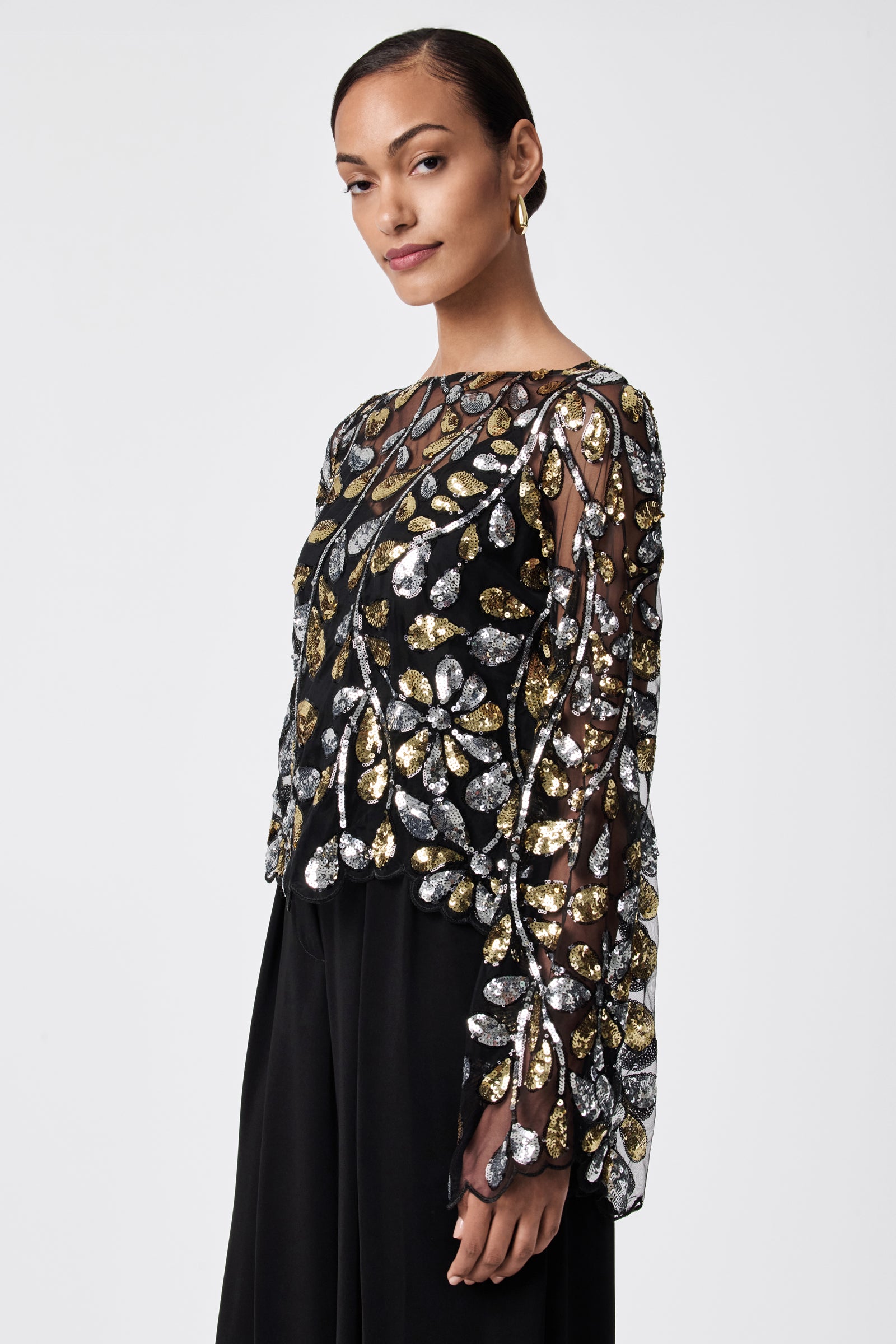 A woman models the FEMMES TOP— a sheer black 100% nylon top with gold and silver sequin floral patterns—styled with black wide-leg pants, slicked-back hair, and hoop earrings against a plain background.