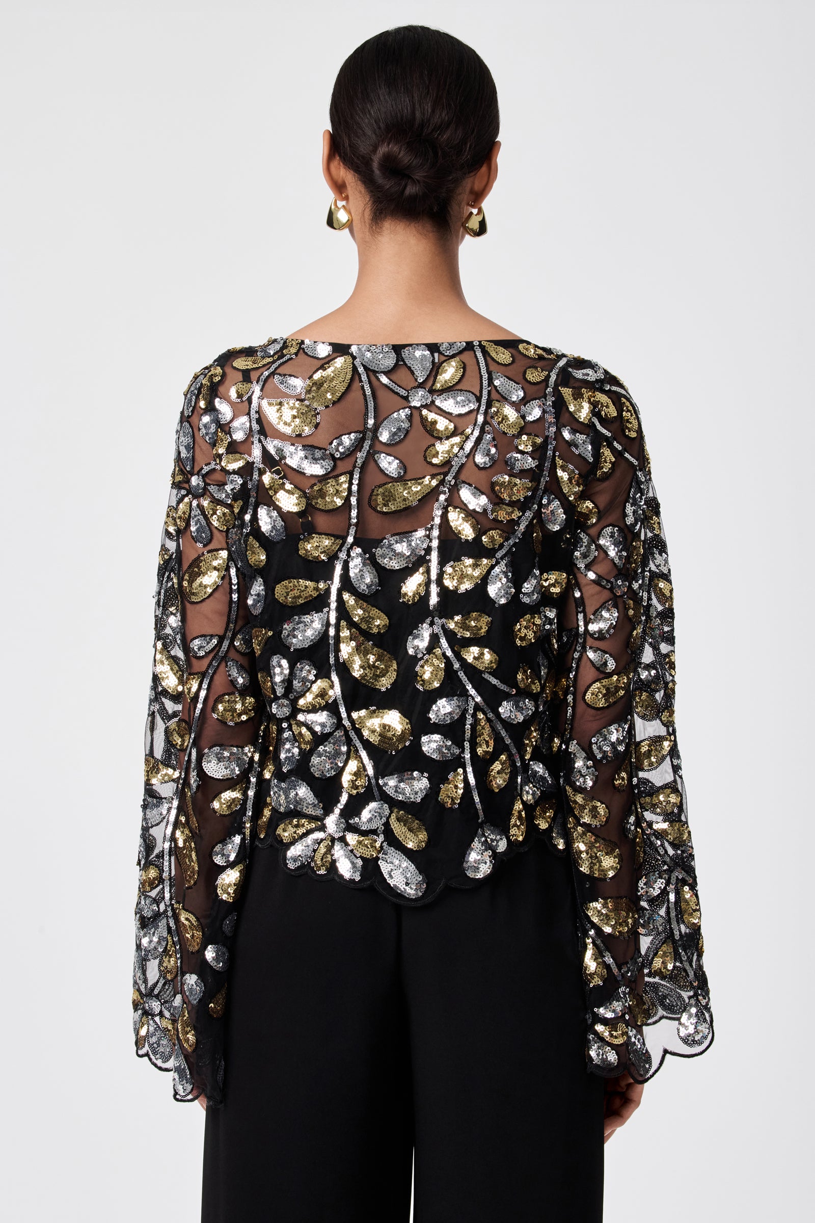 A person with dark hair in a low bun models the FEMMES TOP—a sheer black 100% nylon blouse featuring gold and silver sequin leaf patterns and wide sleeves—paired with black pants, photographed from the back against a plain light background.