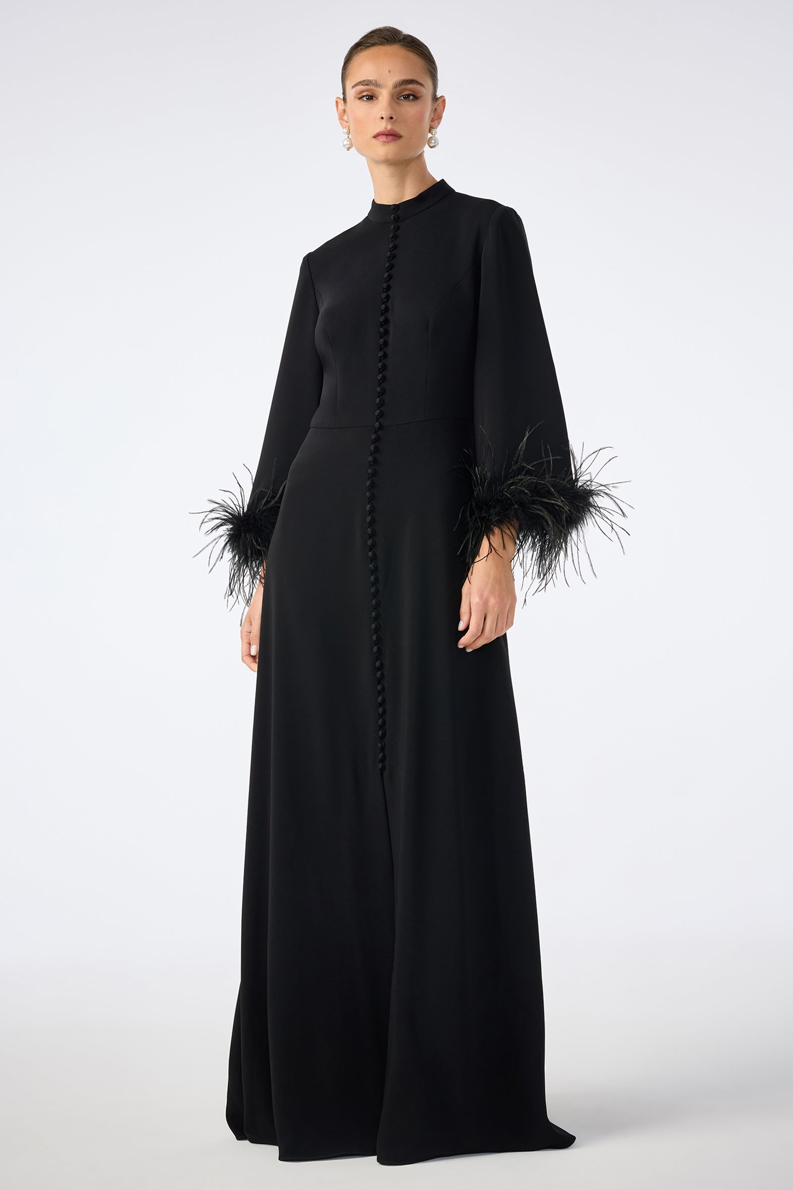 A woman poses against a plain white background wearing the Montmarte Dress—a long black, button-down dress with long sleeves and feathered cuffs.