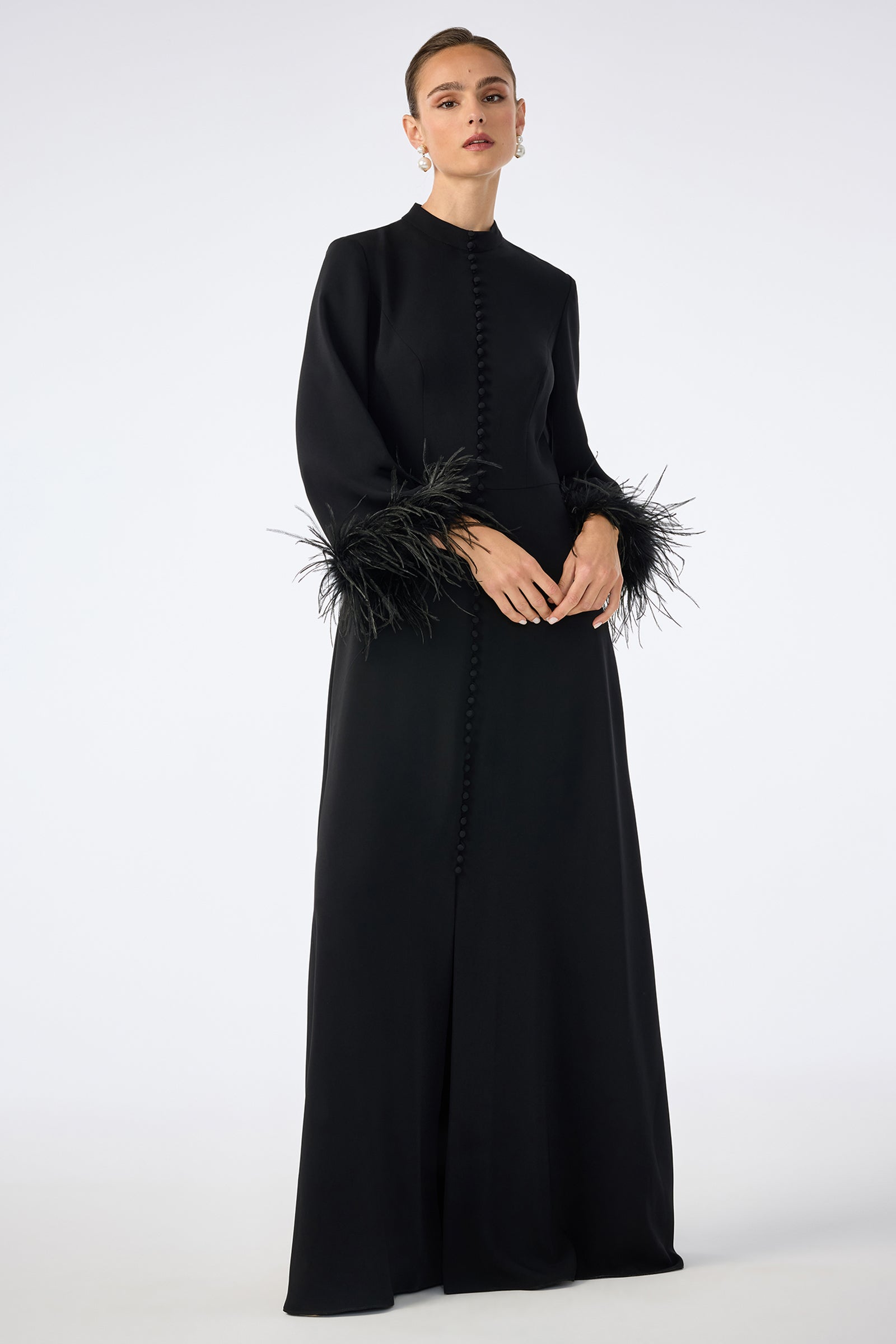 A person wears the Montmarte Dress, a long black gown featuring a high neckline, front button details, and dramatic feathered cuffs, standing against a plain light background.