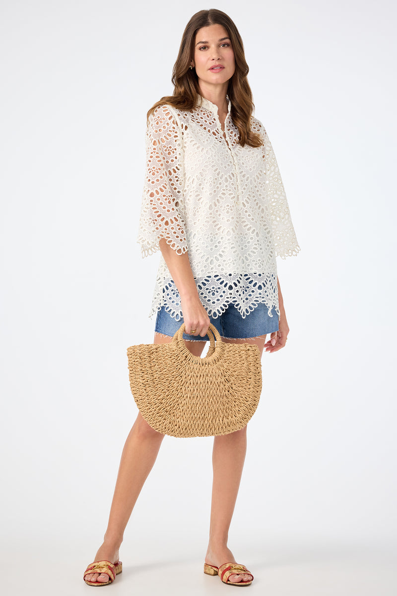 A woman in the Martigues Lace Top, denim shorts, and sandals holds a woven straw handbag while standing against a plain light background.