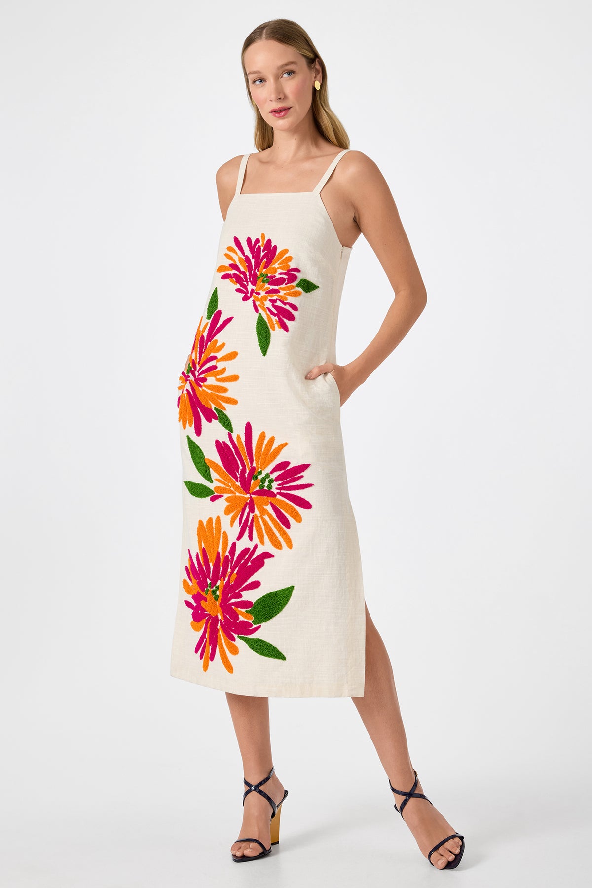 A woman wears the Yona Embroidered Flowers Midi Dress, a sleeveless, light-hued midi with bold pink, orange, and yellow floral prints. She has one hand in her pocket and pairs it with dark strappy heels.