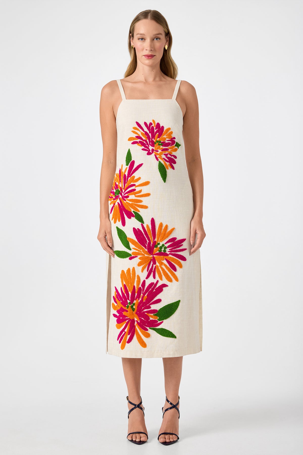 A woman stands facing forward in the Yona Embroidered Flowers Midi Dress‚Äîan ankle-length, sleeveless cream dress with bold pink, orange, and red floral prints. She wears strappy black sandals and has straight hair, set against a plain white background.