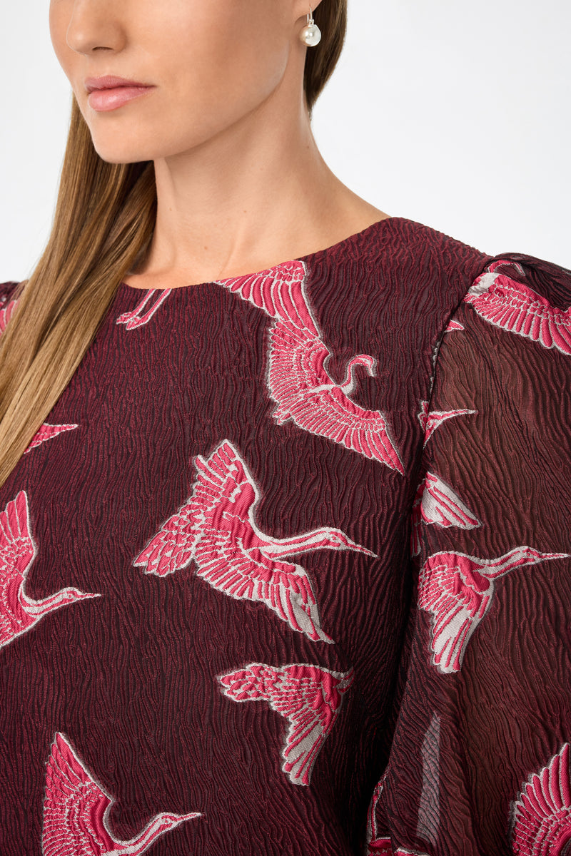 A woman models the Ringo Short Puff Sleeve Mini Dress‚Äîa textured burgundy piece with pink and white crane embroidery. Only her lower face, a single earring, and part of her straight hair are visible.