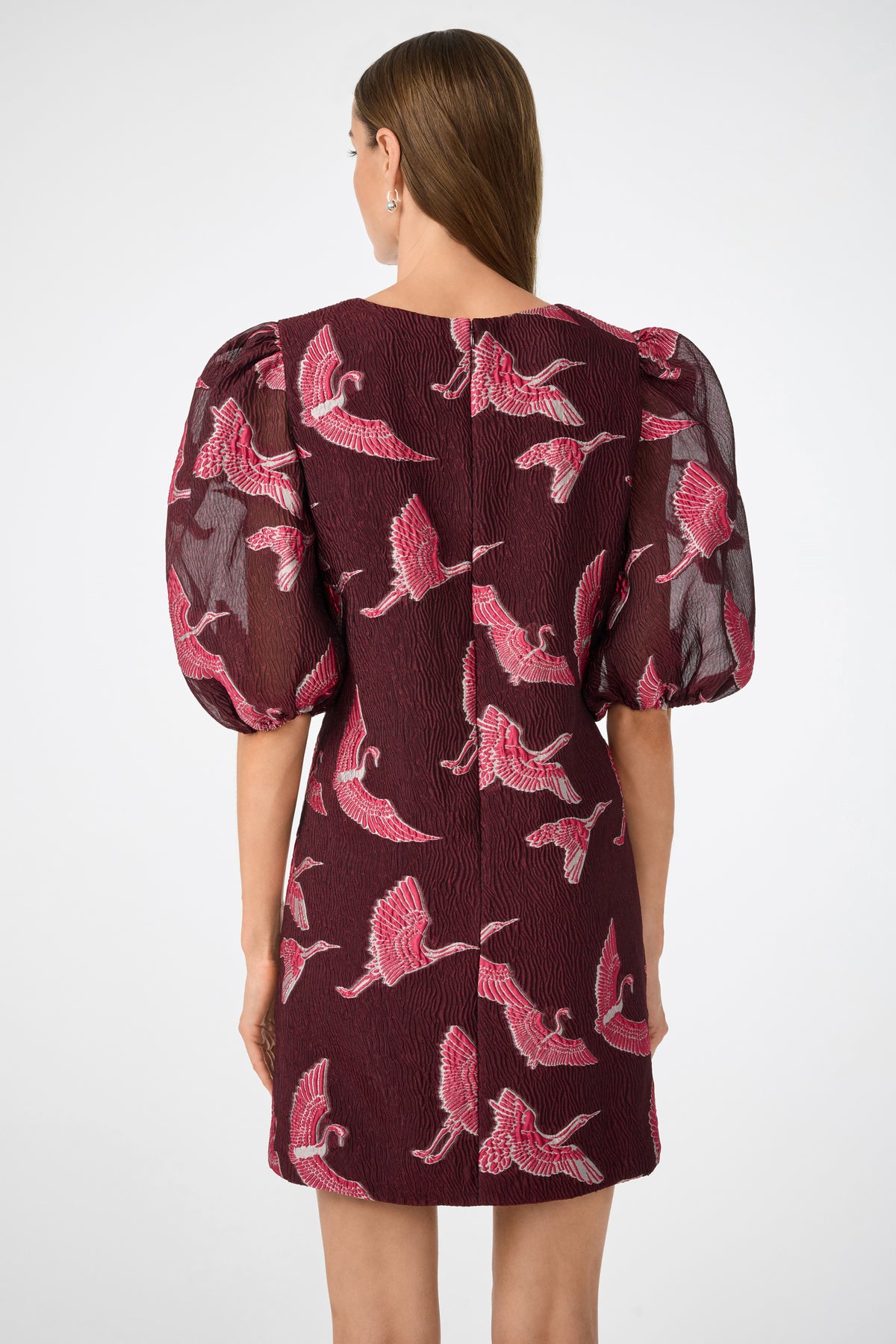 A woman with straight brown hair models the Ringo Short Puff Sleeve Mini Dress in maroon, which has textured fabric, pink and white crane patterns, and puffed mid-length sleeves. She is shown from the back against a plain background.