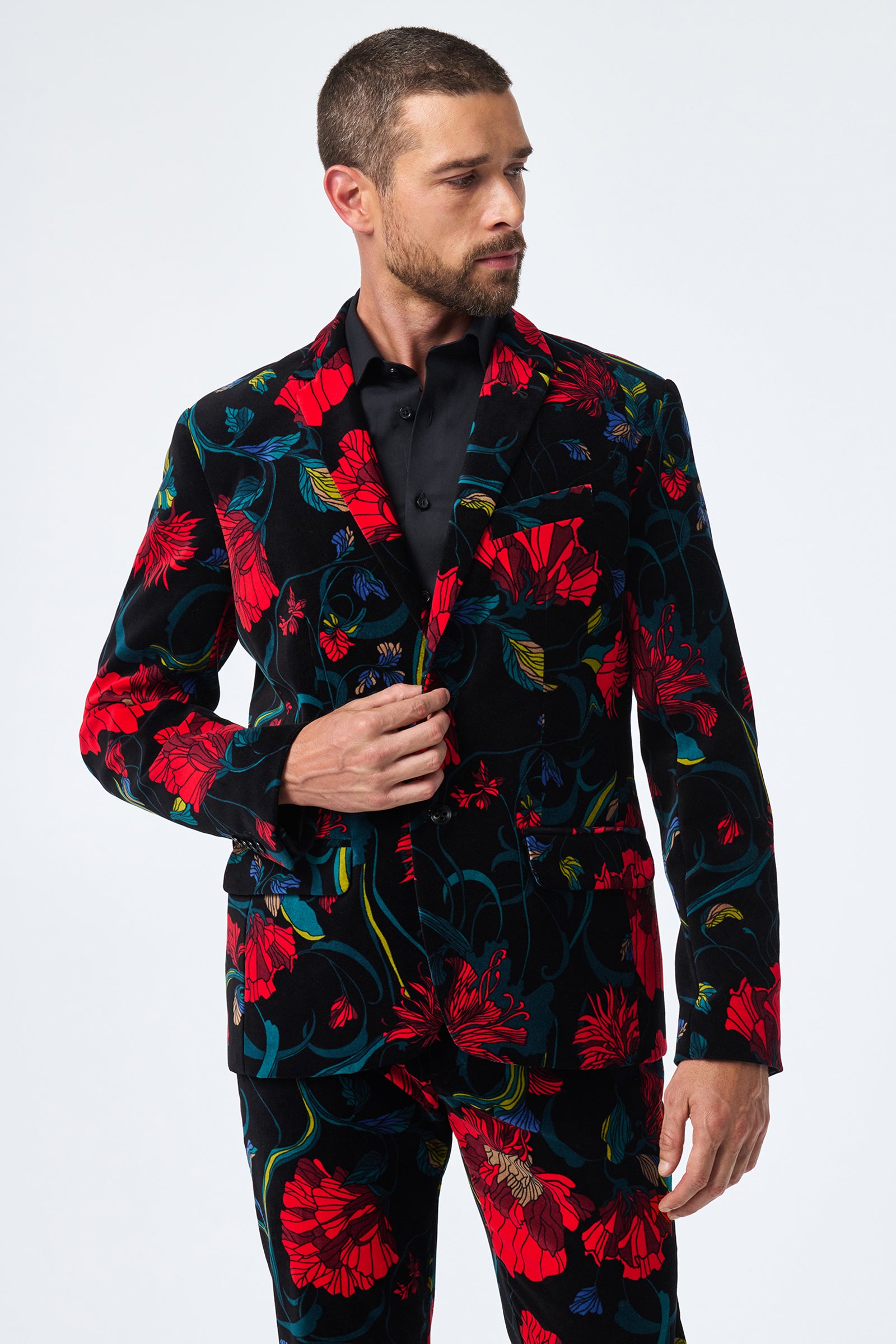 A man with short hair and a beard wears the Thurston Printed Velvet Blazer—black with a bold red and green floral pattern—over a black shirt. He looks to his left, holding the jacket lapel, against a plain white background.