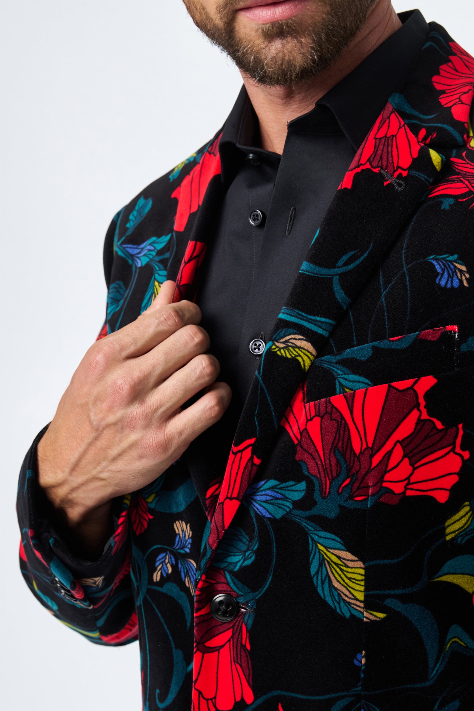A man in the Thurston Printed Velvet Blazer—black with a vibrant red, yellow, and blue floral pattern—adjusts his lapel with his right hand. His black shirt is visible, but his face is only partially shown.