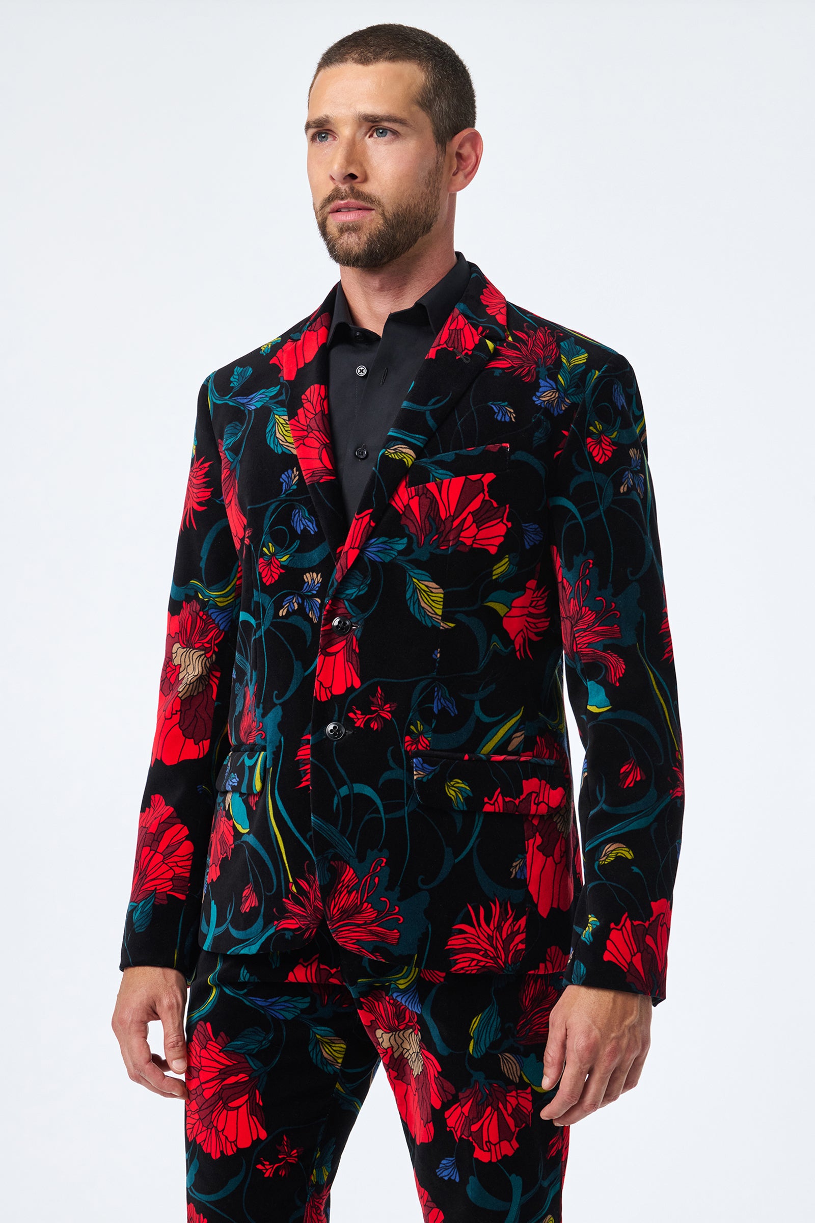 A man wears the Thurston Printed Velvet Blazer, featuring a striking black base with red, blue, and green floral patterns, paired with a black dress shirt. He looks slightly to the side against a plain white background.