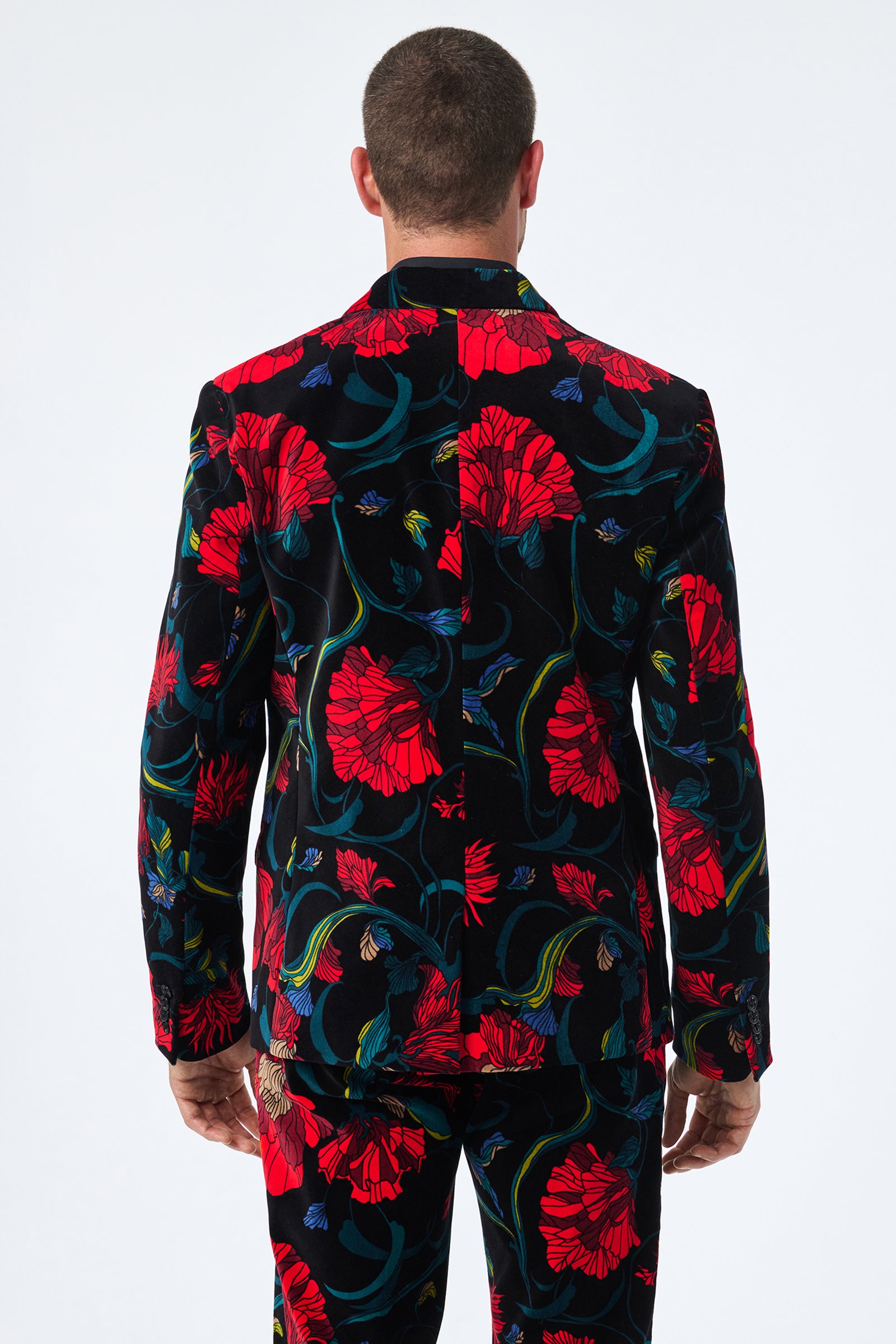 A man is seen from behind in the Thurston Printed Velvet Blazer, featuring a black base with vibrant red floral patterns and green and blue accents; he has short hair and stands with relaxed arms at his sides.