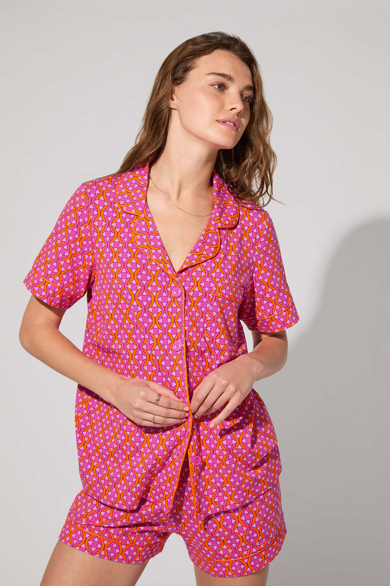 A person wears the Shorty PJ Set - Chain Geo, a matching short-sleeve button-up shirt and shorts featuring a vibrant pink and orange geometric pattern, standing against a plain light background.