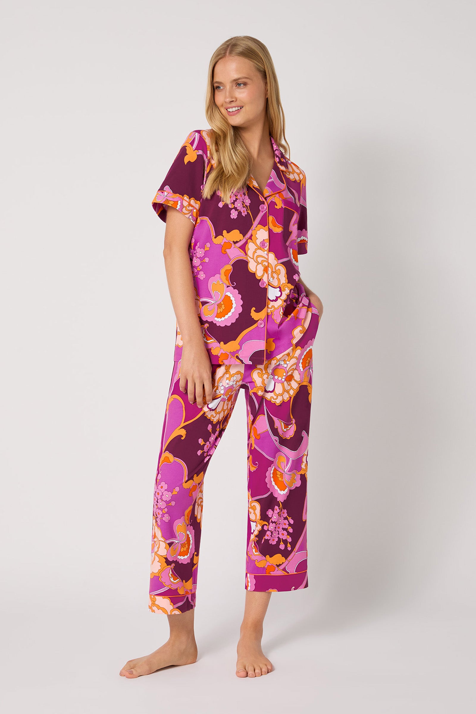 A woman stands barefoot against a plain background, smiling with one hand in her pocket, wearing the Apache Plume S/S Cropped PJ: a vibrant matching pajama set featuring a bold pink, purple, orange, and white floral pattern and short sleeves.