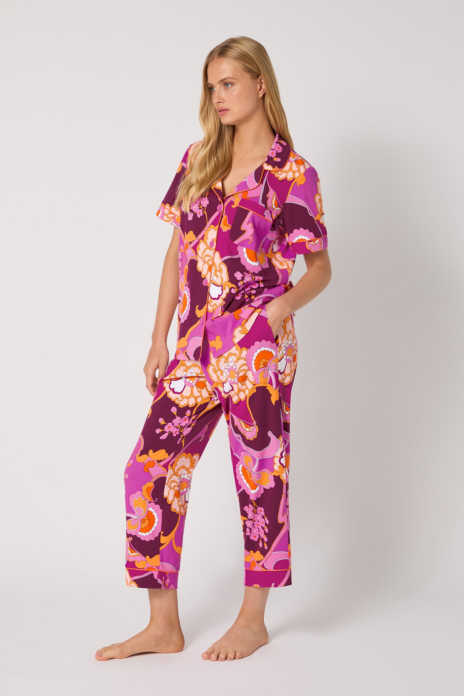 A woman stands barefoot in the Apache Plume S/S Cropped Pj, a matching set with vibrant pink, purple, and orange floral patterns, featuring short sleeves and cropped pants against a plain light background.