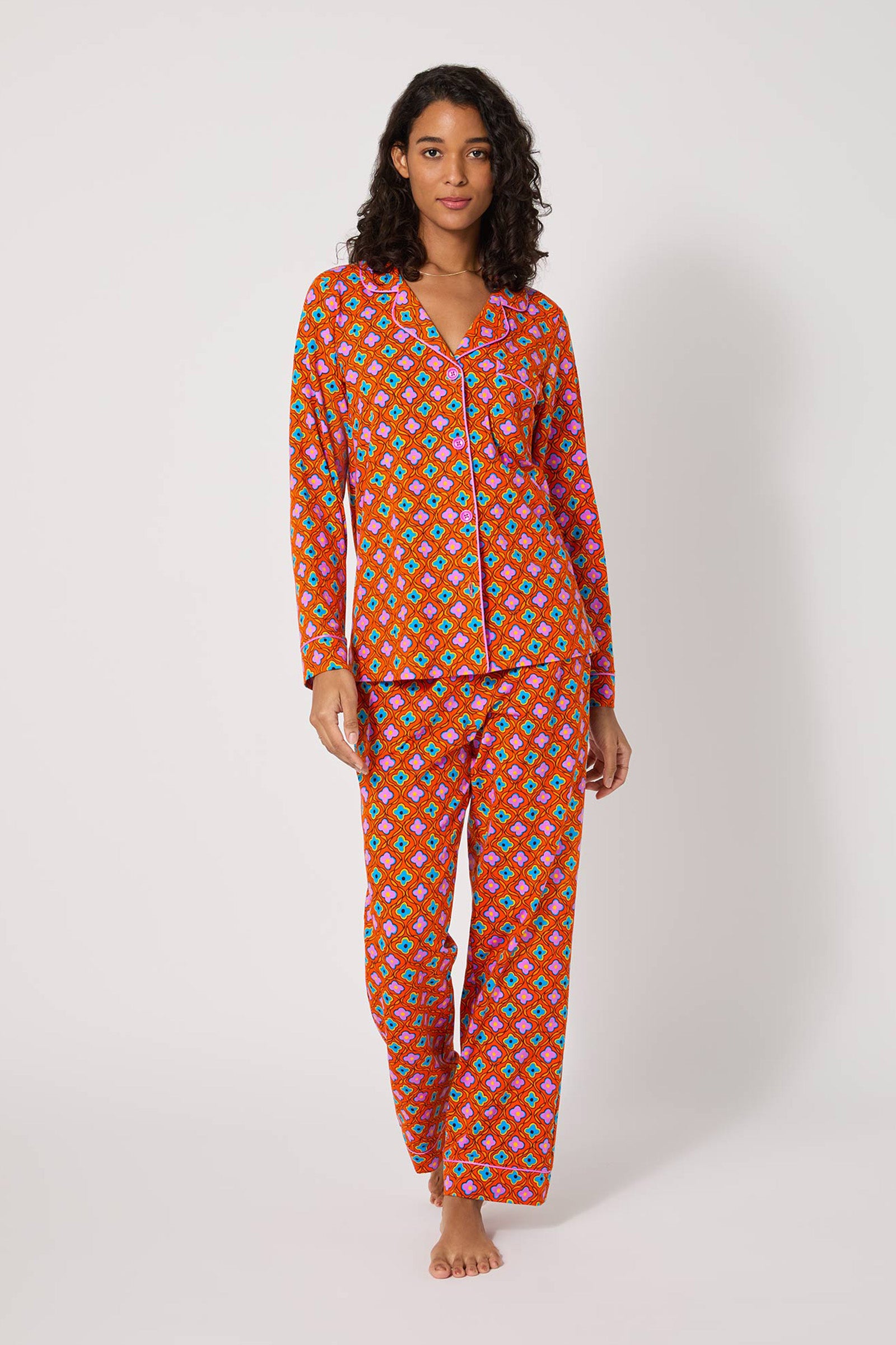 A woman stands barefoot against a plain background, wearing the Daisy Foulard L/S Classic Pj—a red long-sleeve, button-up pajama set featuring a colorful geometric print.