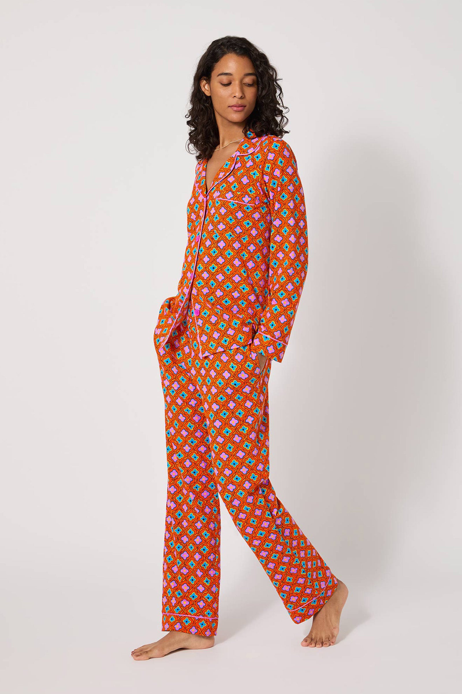 A woman stands barefoot against a plain background, wearing the Daisy Foulard L/S Classic Pj—a bright red set with a button-up top and long pants decorated in small colorful patterns. She has her hands in her pockets and looks relaxed.
