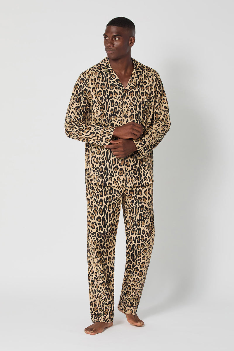 A man with short hair stands barefoot against a white background, wearing the Leopard Stripe Mens Classic Pj set featuring a button-up shirt and long pants in matching leopard print. He looks to the side with his hands loosely clasped.