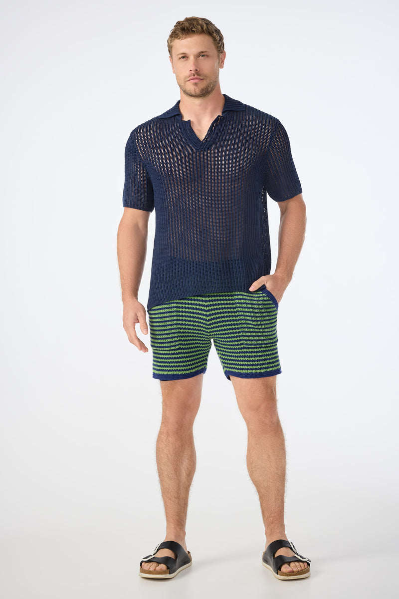A man poses against a plain background wearing the Javi Crochet Pullover - Noh Nightfall, paired with green and navy striped shorts and black sandals. He stands with one hand in his pocket and a neutral expression.