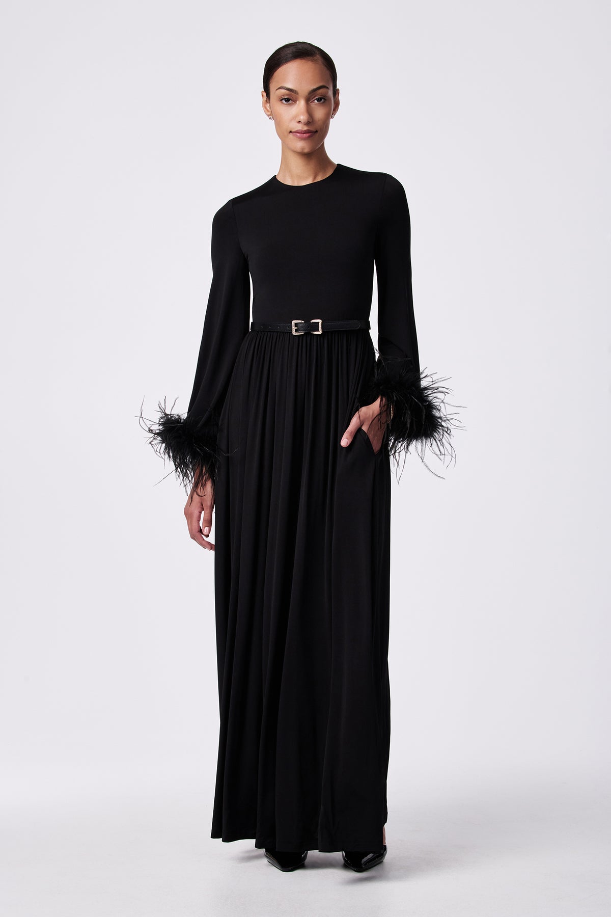 A woman poses against a white background in the Orla Feather-Cuff Maxi Dress, featuring a belted waist and feathered sleeve cuffs. Her hair is pulled back and one hand rests in her pocket.
