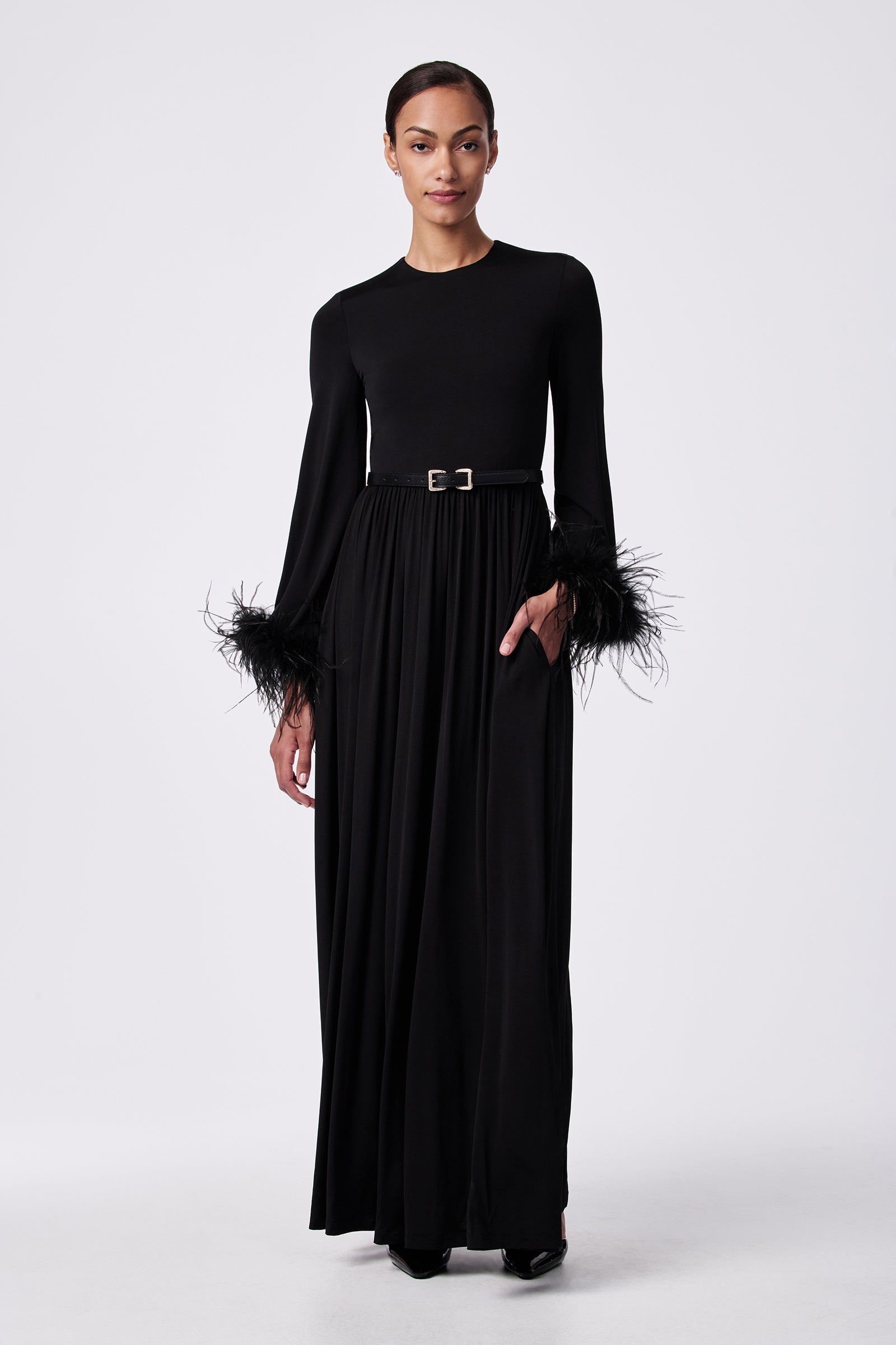A woman poses against a white background in the Orla Feather-Cuff Maxi Dress, featuring a belted waist and feathered sleeve cuffs. Her hair is pulled back and one hand rests in her pocket.