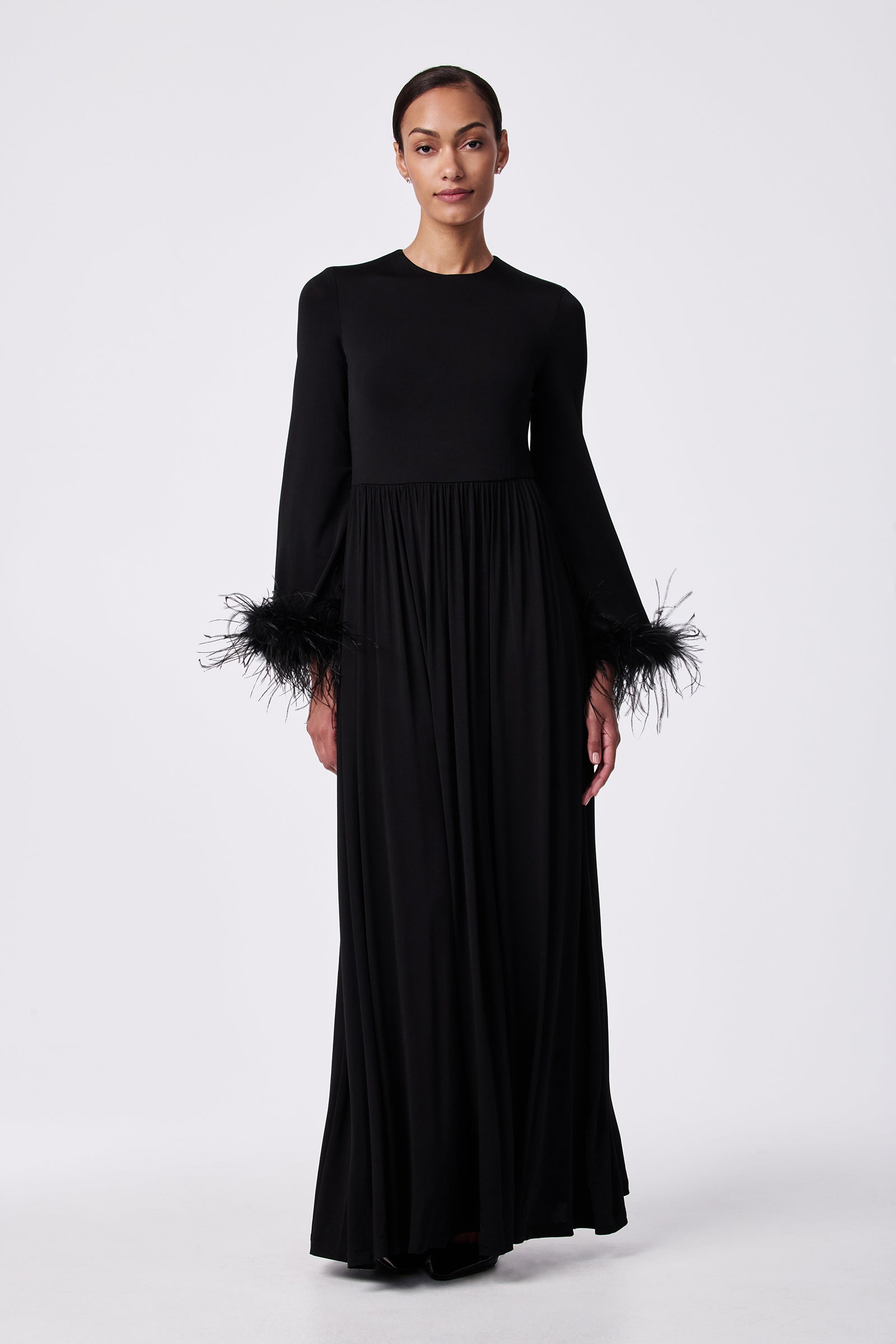 A woman wears the Orla Feather-Cuff Maxi Dress, featuring a fitted bodice, round neckline, long sleeves with black feather cuffs, and a floor-length skirt against a plain white background.