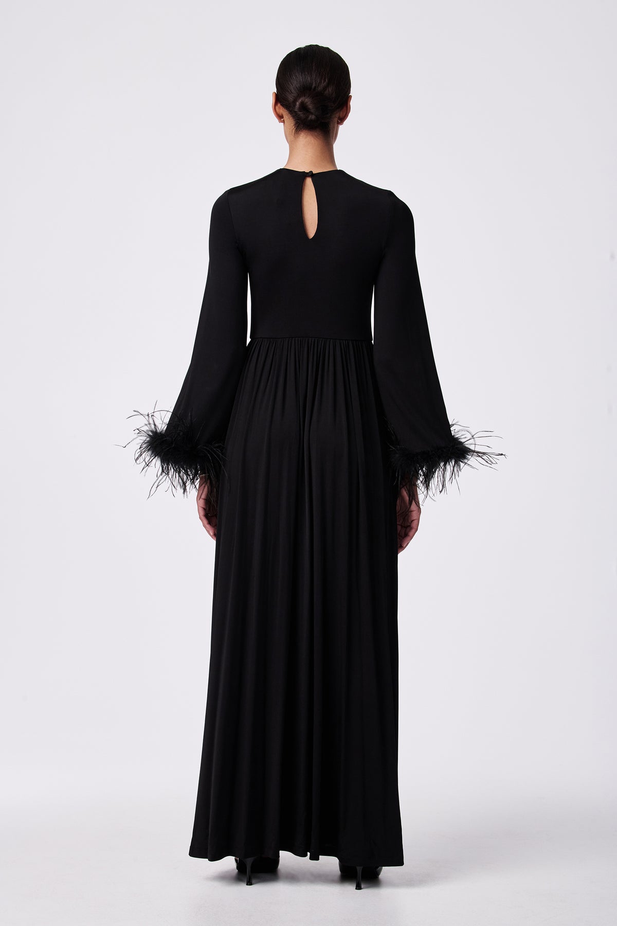 A person stands facing away, wearing the Orla Feather-Cuff Maxi Dressâ€”a long black gown with flared feather-trimmed sleeves, a small keyhole opening at the upper back, and hair styled in a low bun.