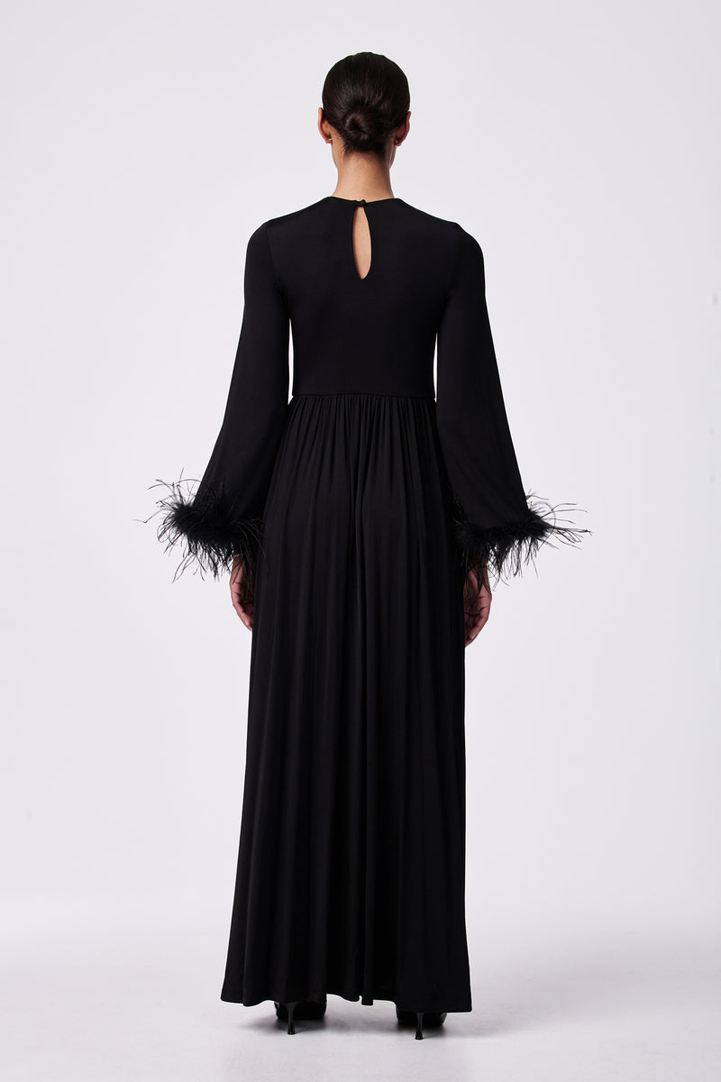 A person stands facing away, wearing the Orla Feather-Cuff Maxi Dressâ€”a long black gown with flared feather-trimmed sleeves, a small keyhole opening at the upper back, and hair styled in a low bun.