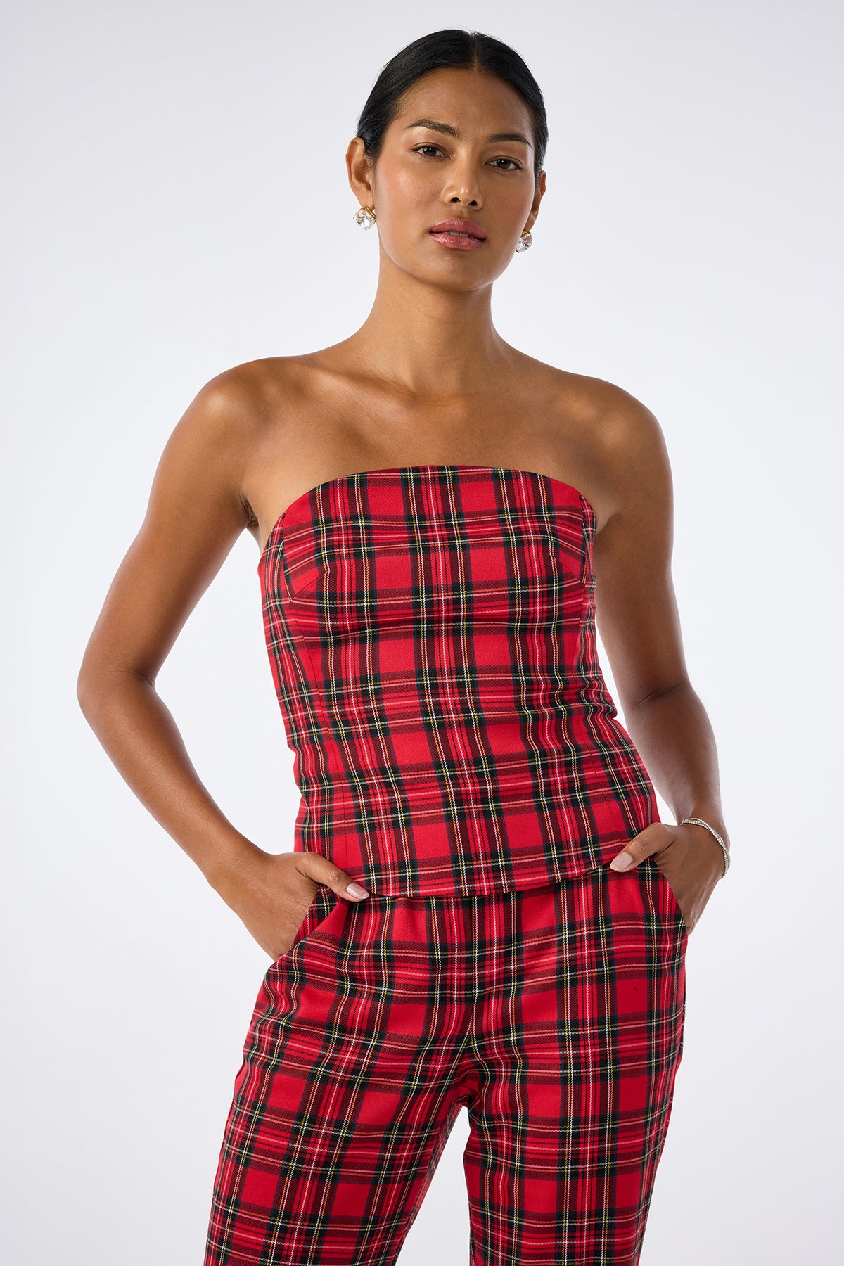 A woman stands against a plain background wearing the Arden Top, a red plaid strapless piece, paired with matching pants. She has her hands in her pockets and maintains a neutral expression.