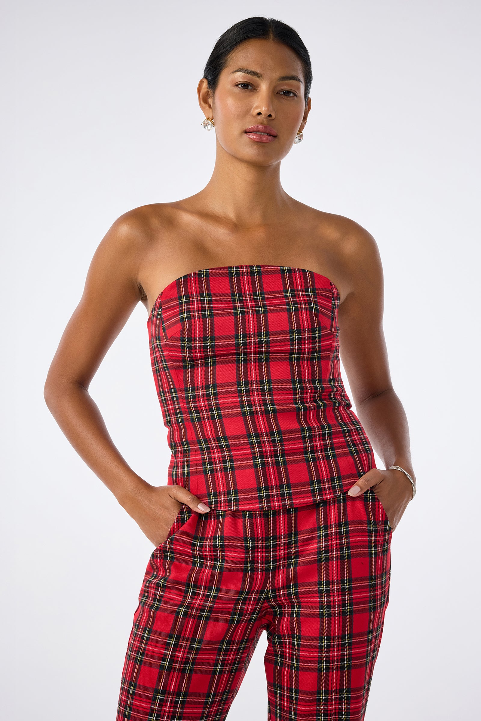 A woman stands against a plain background wearing the Arden Top, a red plaid strapless piece, paired with matching pants. She has her hands in her pockets and maintains a neutral expression.
