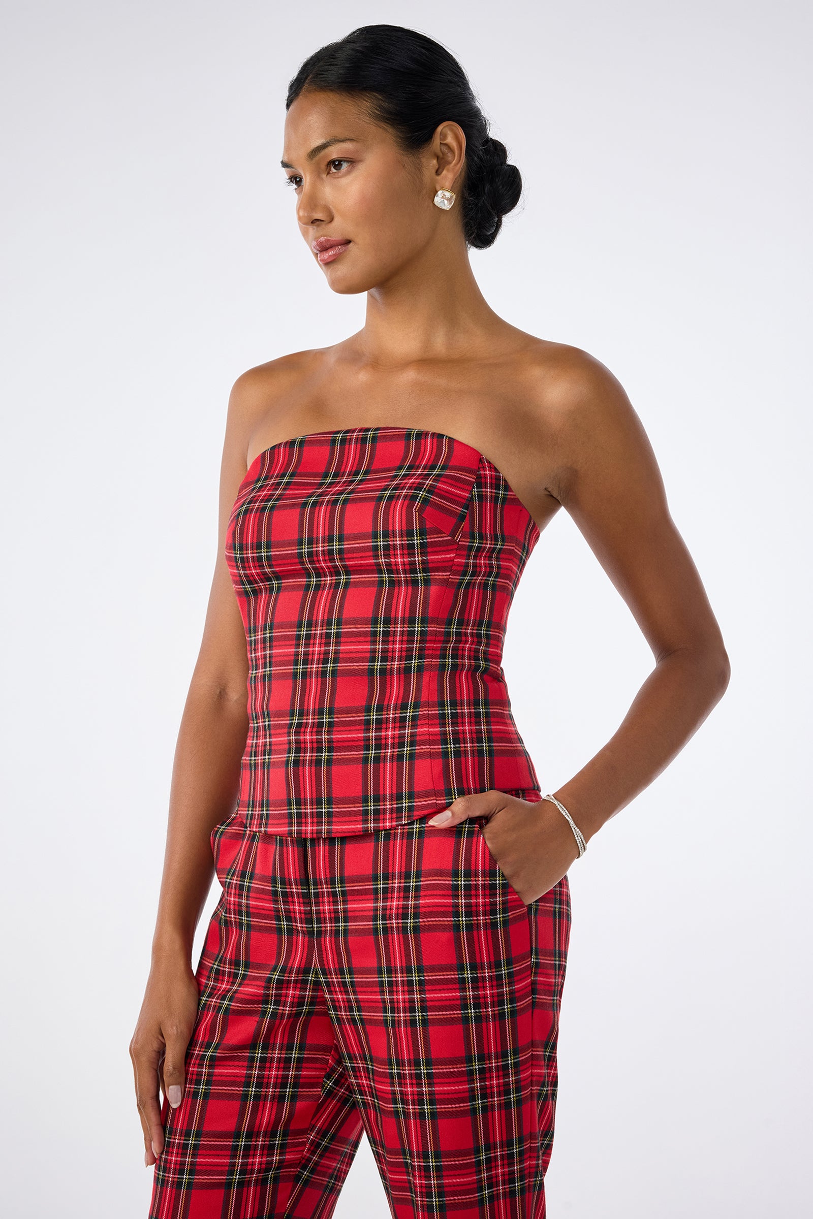 A woman wears the Arden Top, a red plaid strapless piece, paired with matching pants. She stands with one hand in her pocket, looking to the side, her hair in a low bun, accessorized with earrings and a bracelet.