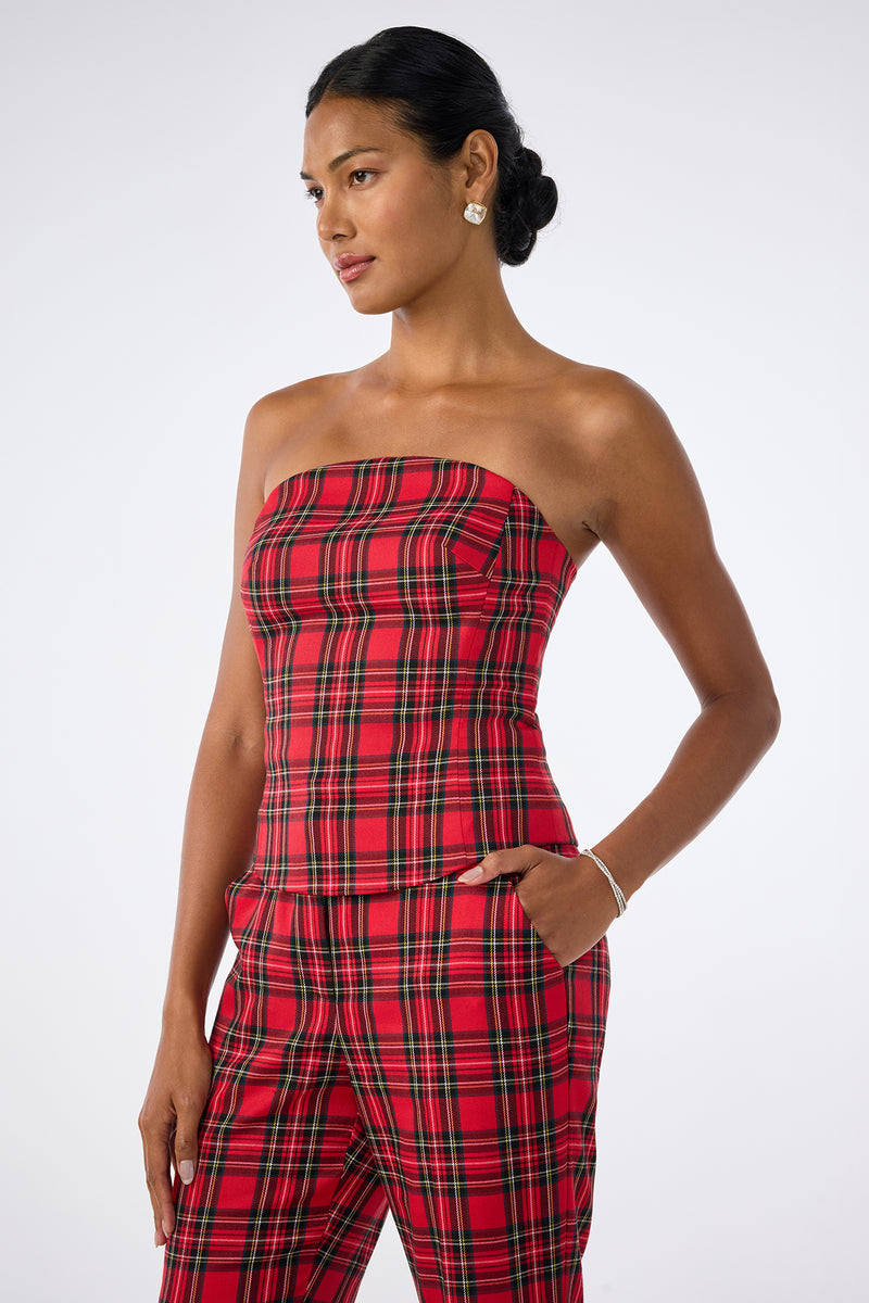 A woman wears the Arden Top, a red plaid strapless piece, paired with matching pants. She stands with one hand in her pocket, looking to the side, her hair in a low bun, accessorized with earrings and a bracelet.