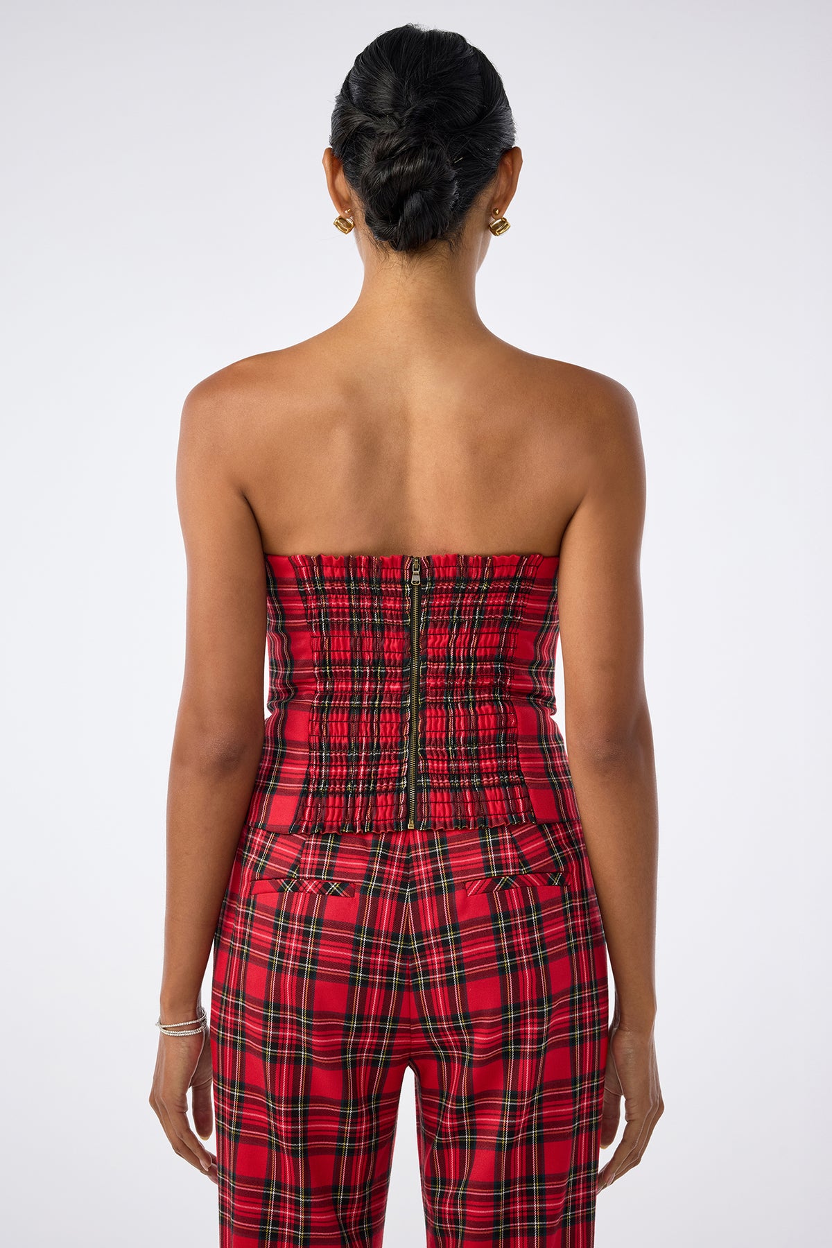A person is seen from behind wearing the strapless Arden Top in red plaid with matching pants. Their hair is styled in a neat bun, accessorized with bracelets on the left wrist and small hoop earrings against a plain white background.