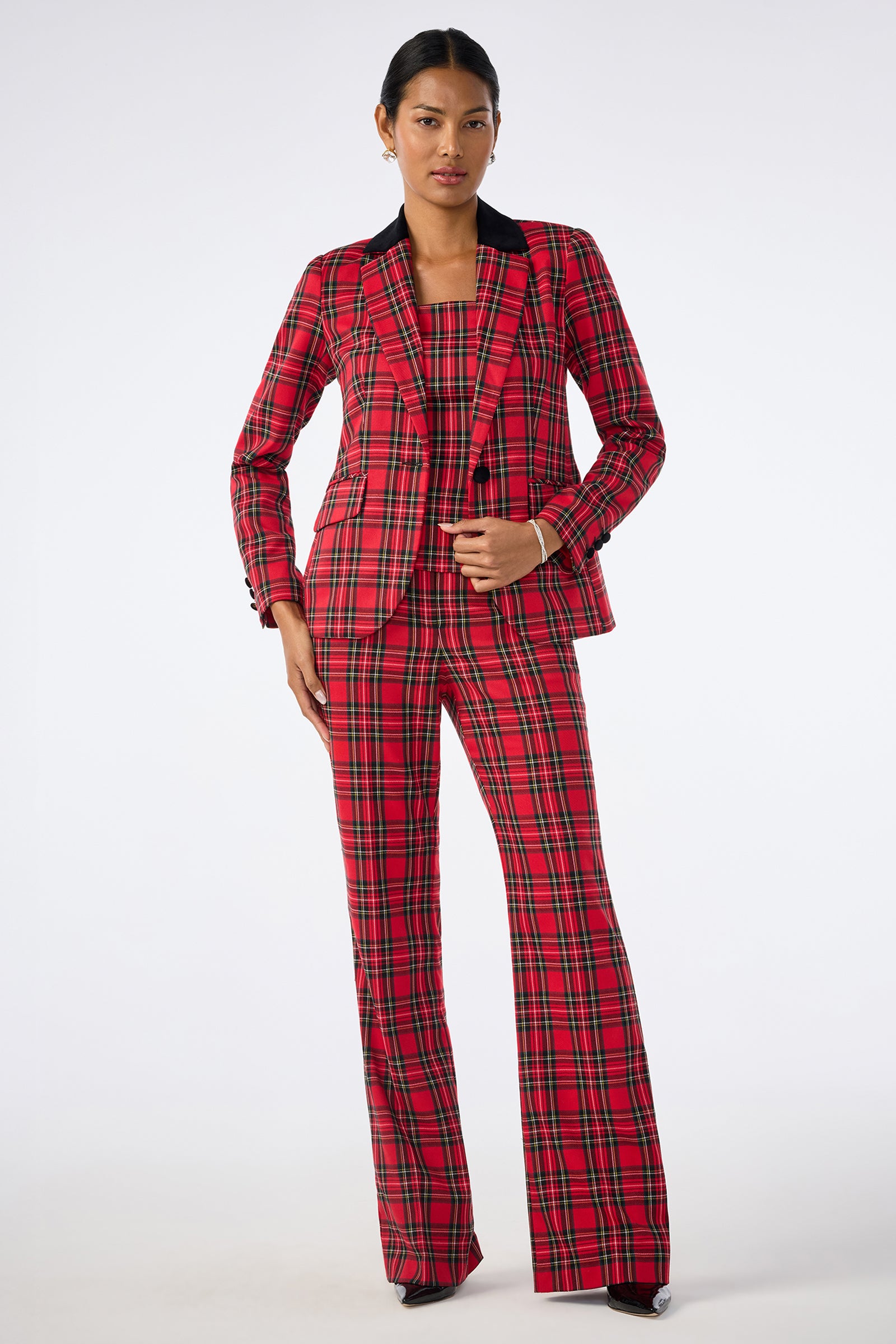 A person poses confidently in the Revolution Jacket, a red plaid blazer with matching flared pants and top. They wear hoop earrings, have short hair and black shoes, standing against a plain white background.
