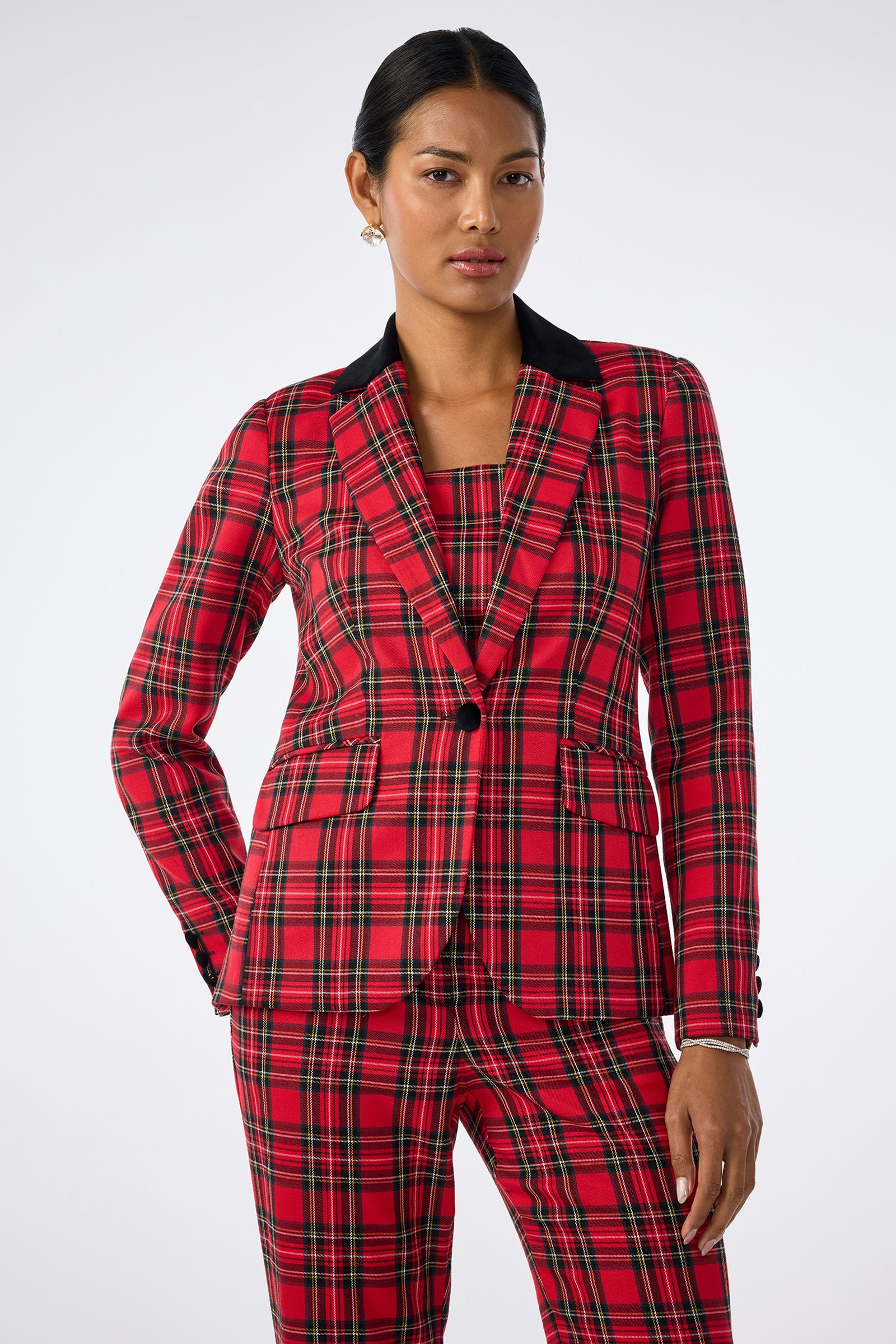 A woman stands against a plain background wearing the Revolution Jacket, a red plaid single-button blazer with a black collar, paired with matching pants. Her hair is pulled back and she wears small earrings.