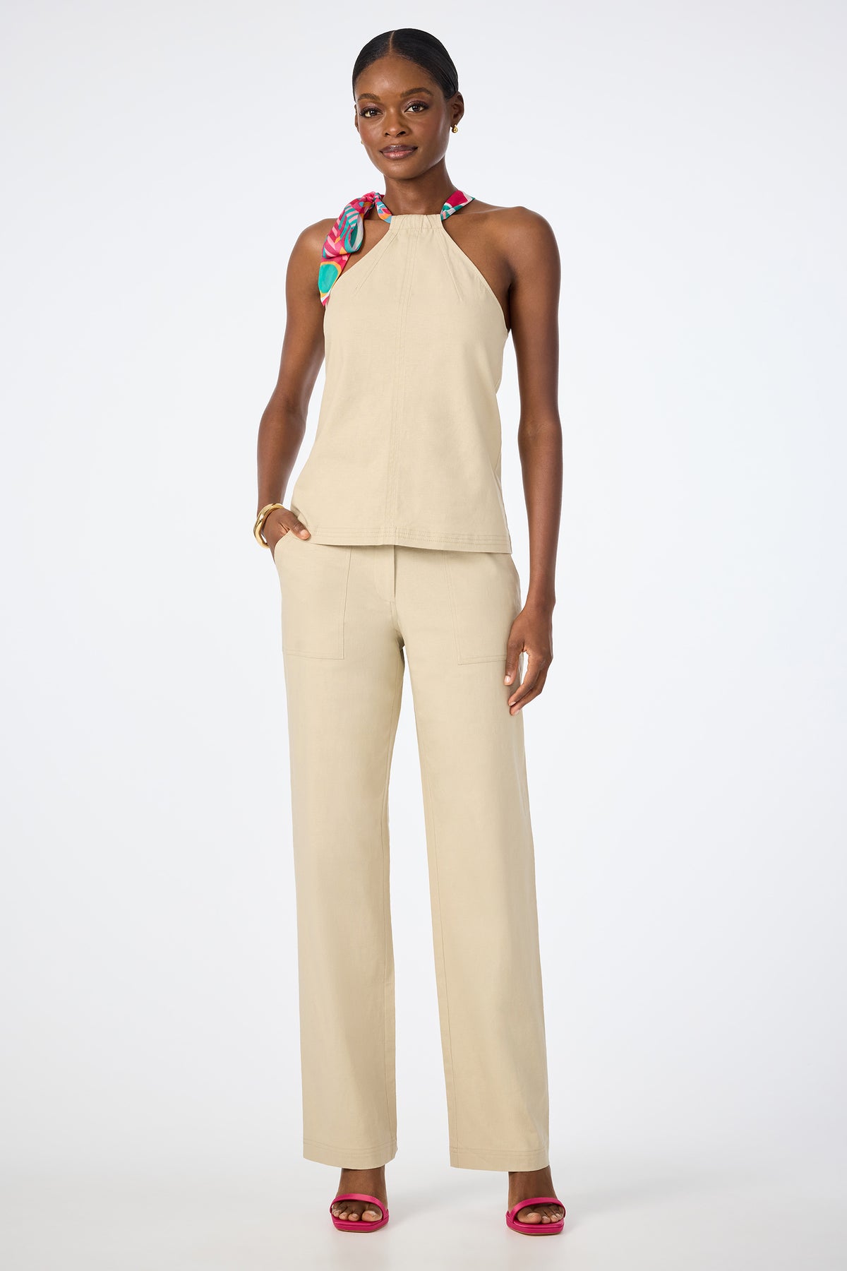 A woman poses against a plain white background wearing beige Antonia 2 Straight Leg Pants and a matching sleeveless halter top, accessorized with a colorful neck scarf and pink high-heeled sandals.