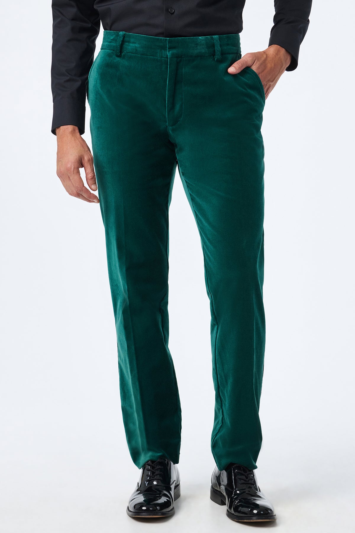 Man wearing Clyde Slim Velvet Trousers in dark green, a black dress shirt, and shiny black dress shoes, with one hand in his pocket against a plain white background.