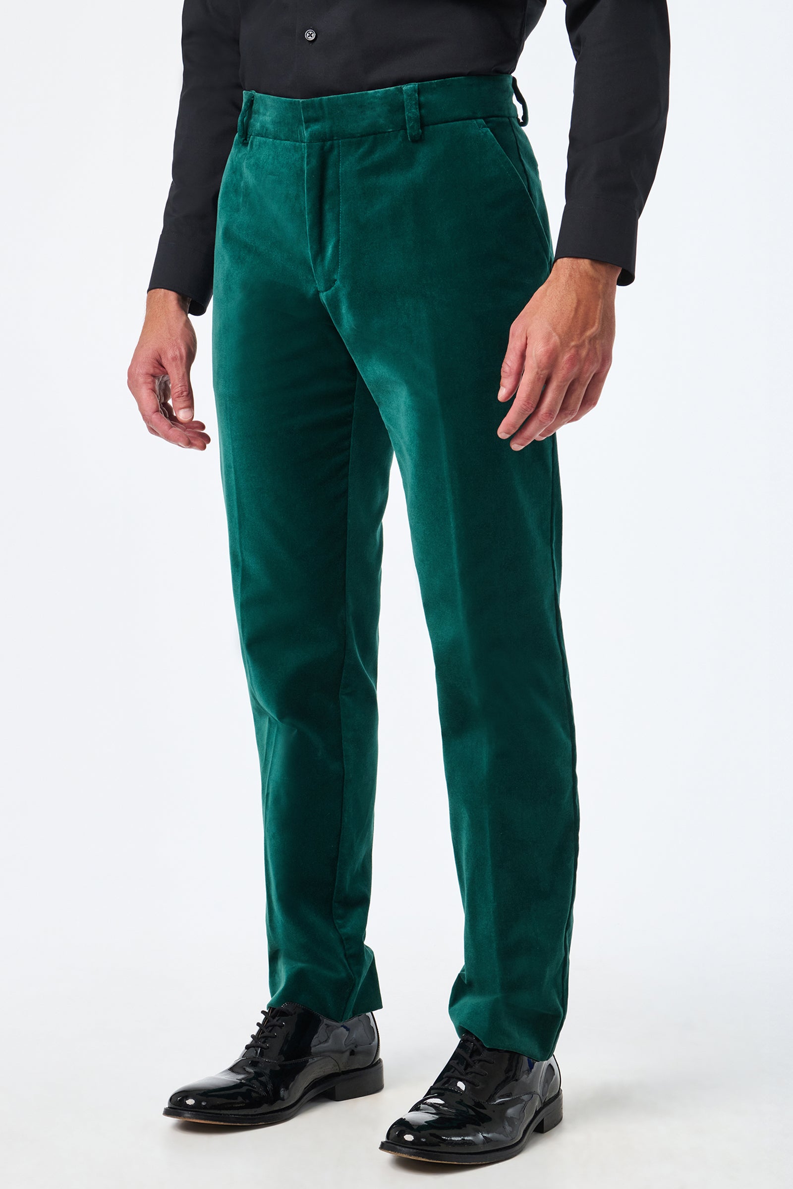 A person wears green Clyde Slim Velvet Trousers, a black dress shirt, and black shiny dress shoes while standing against a plain white background.