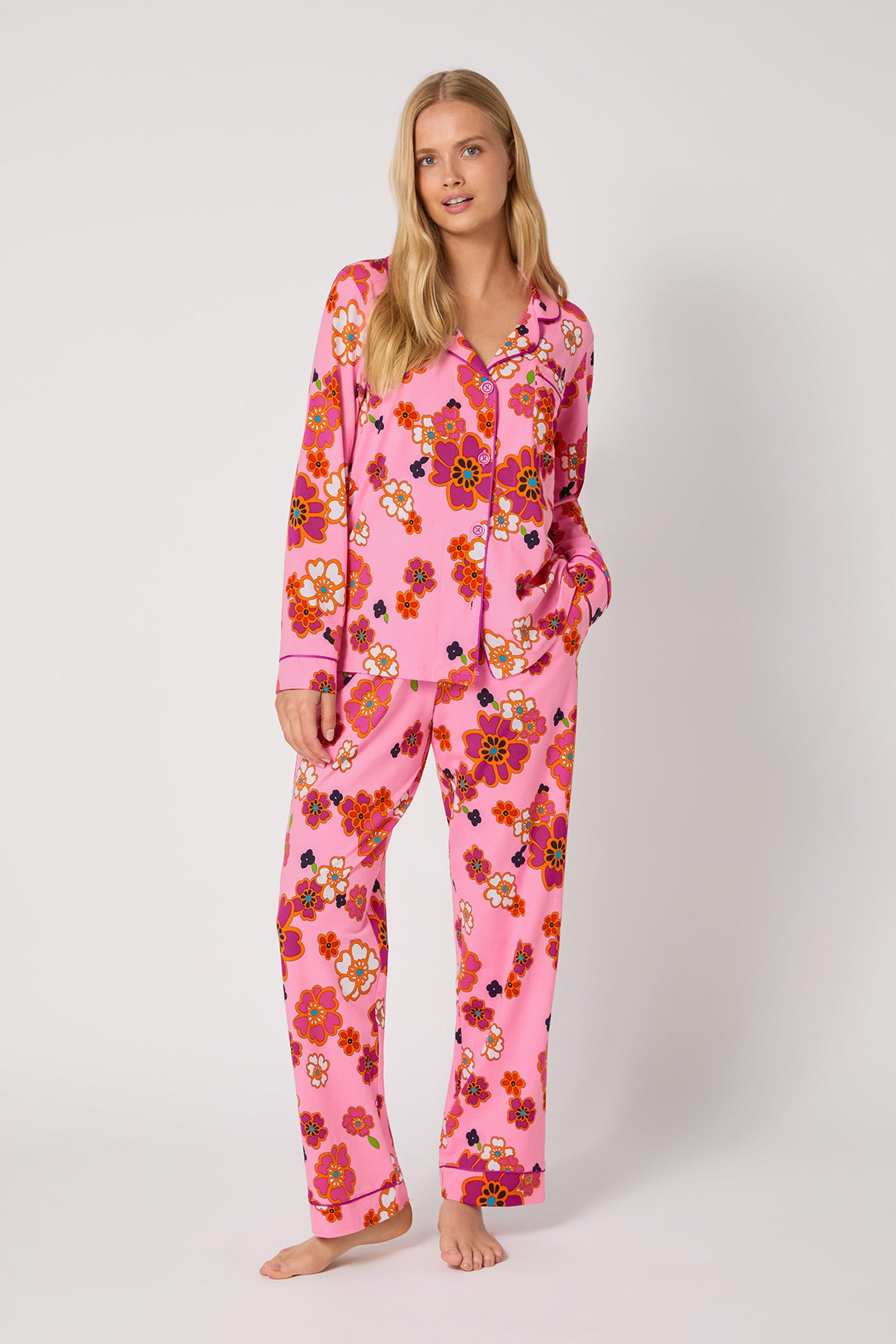 A woman stands barefoot against a plain background, wearing the Resort Floral L/S Classic Pj—a matching pink pajama set with a colorful floral pattern, button-up top with a collar, and one hand in her pocket.
