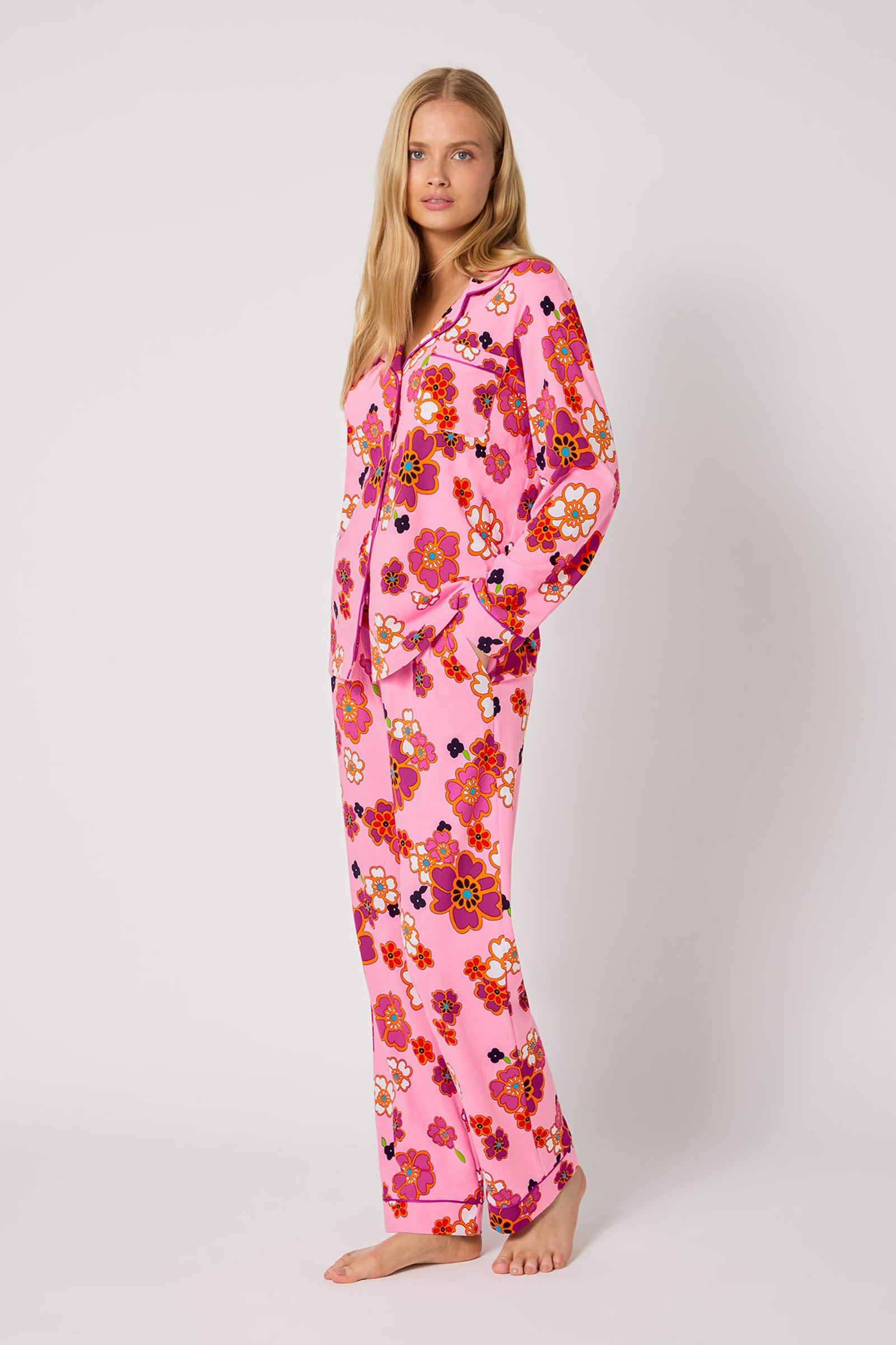 A woman stands barefoot against a plain background, wearing the Resort Floral L/S Classic Pj—a matching pink pajama set with a bold floral pattern, featuring a long-sleeve button-up top and long pants.