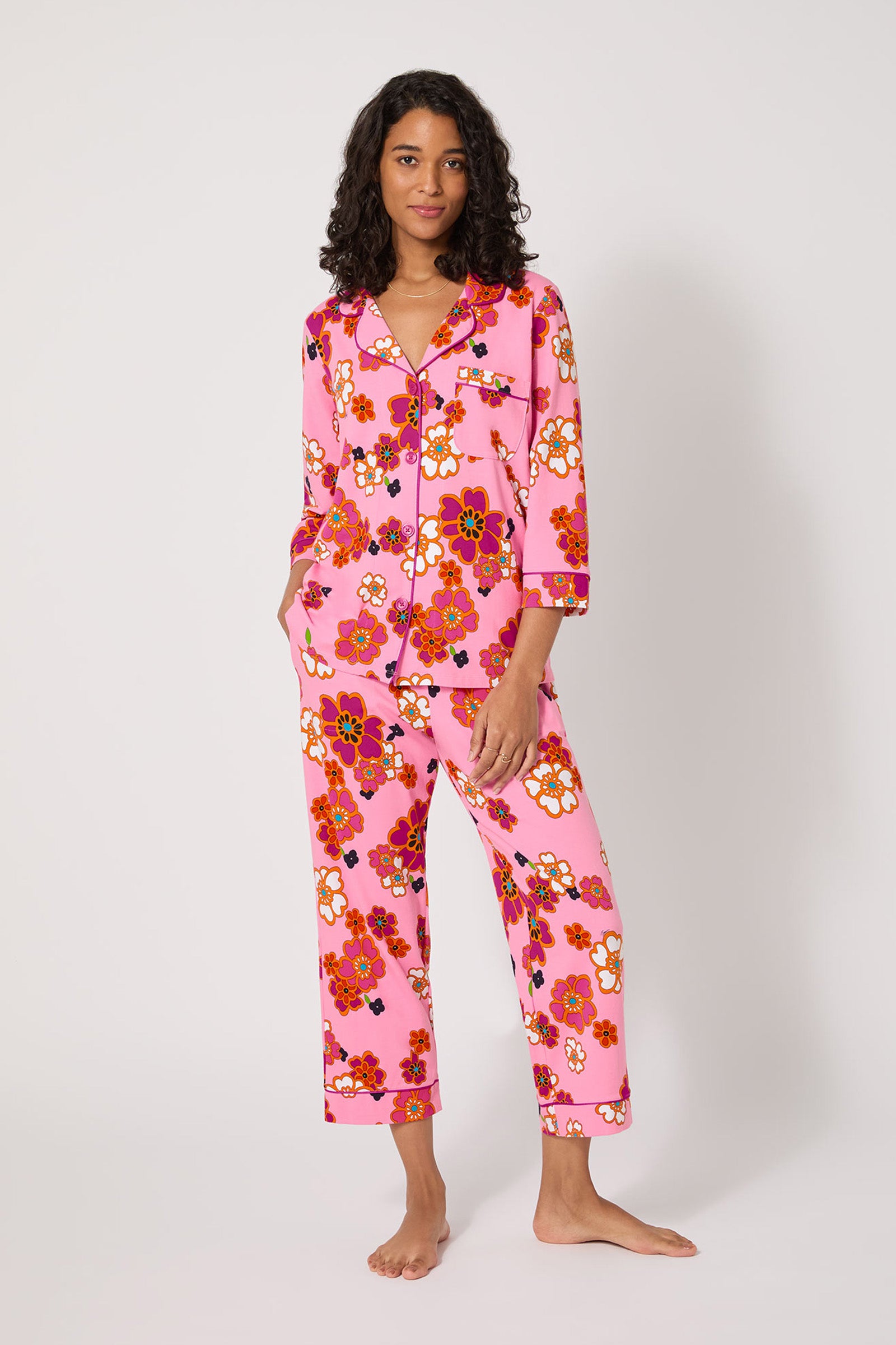 A woman stands barefoot against a plain background, wearing the Resort Floral 3/4 Cropped Pj—pink pajamas with a bold, colorful floral print, 3/4-length sleeves, a collar, full-length pants, hands in her pockets and smiling slightly.