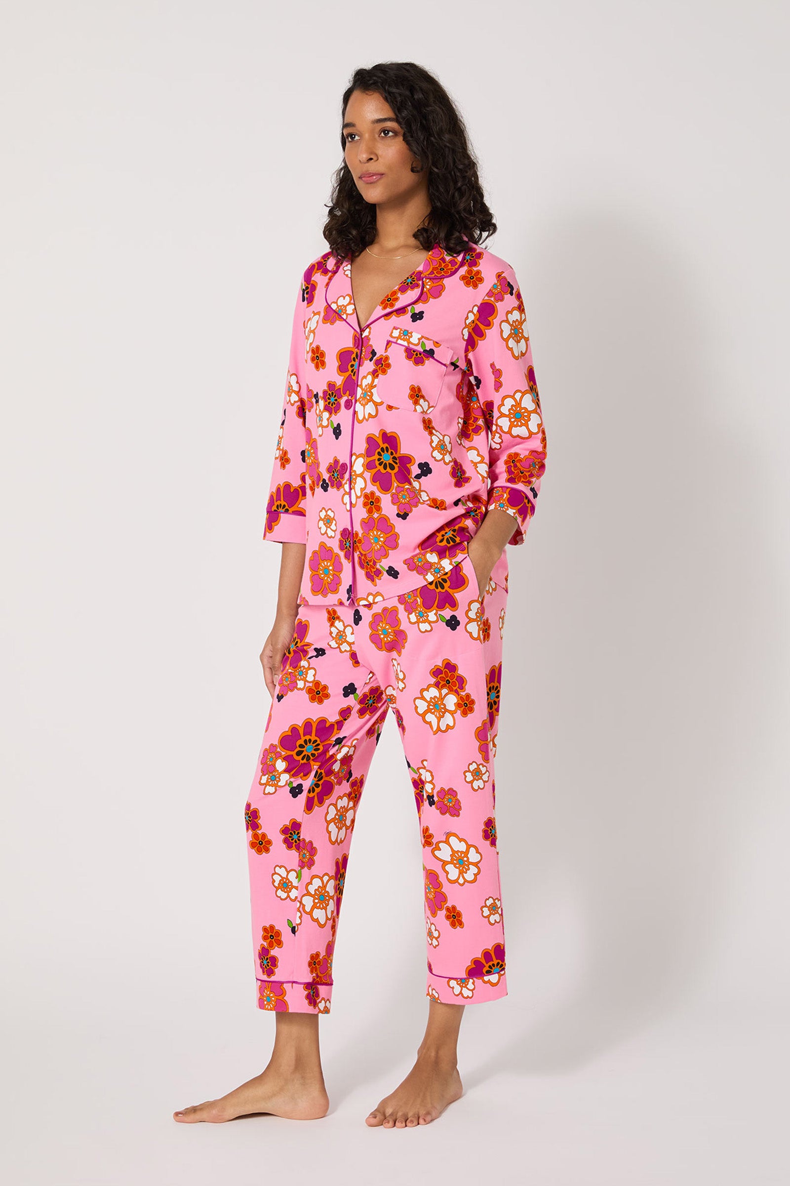 A woman stands barefoot in Resort Floral 3/4 Cropped Pj, featuring pink, red, orange, and white floral print. The pajama set has long sleeves, cropped pants, a button-up front, and is shown against a plain white background.