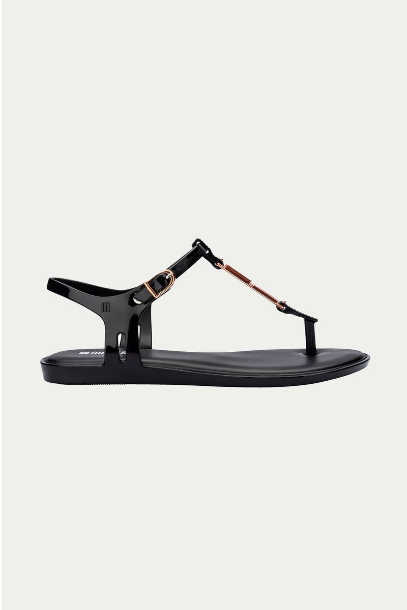 The Melissa Solar Chic T-Strap Sandal in black features a thong design, ankle strap with metallic gold buckle, and metal accents, shown against a plain light background.