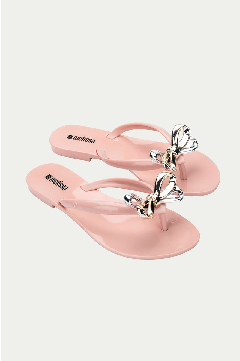 The Melissa Harmonic Eclipse Bow Sandal in pink is a pair of light pink thong sandals featuring metallic butterfly accents on the straps, shown against a white background.