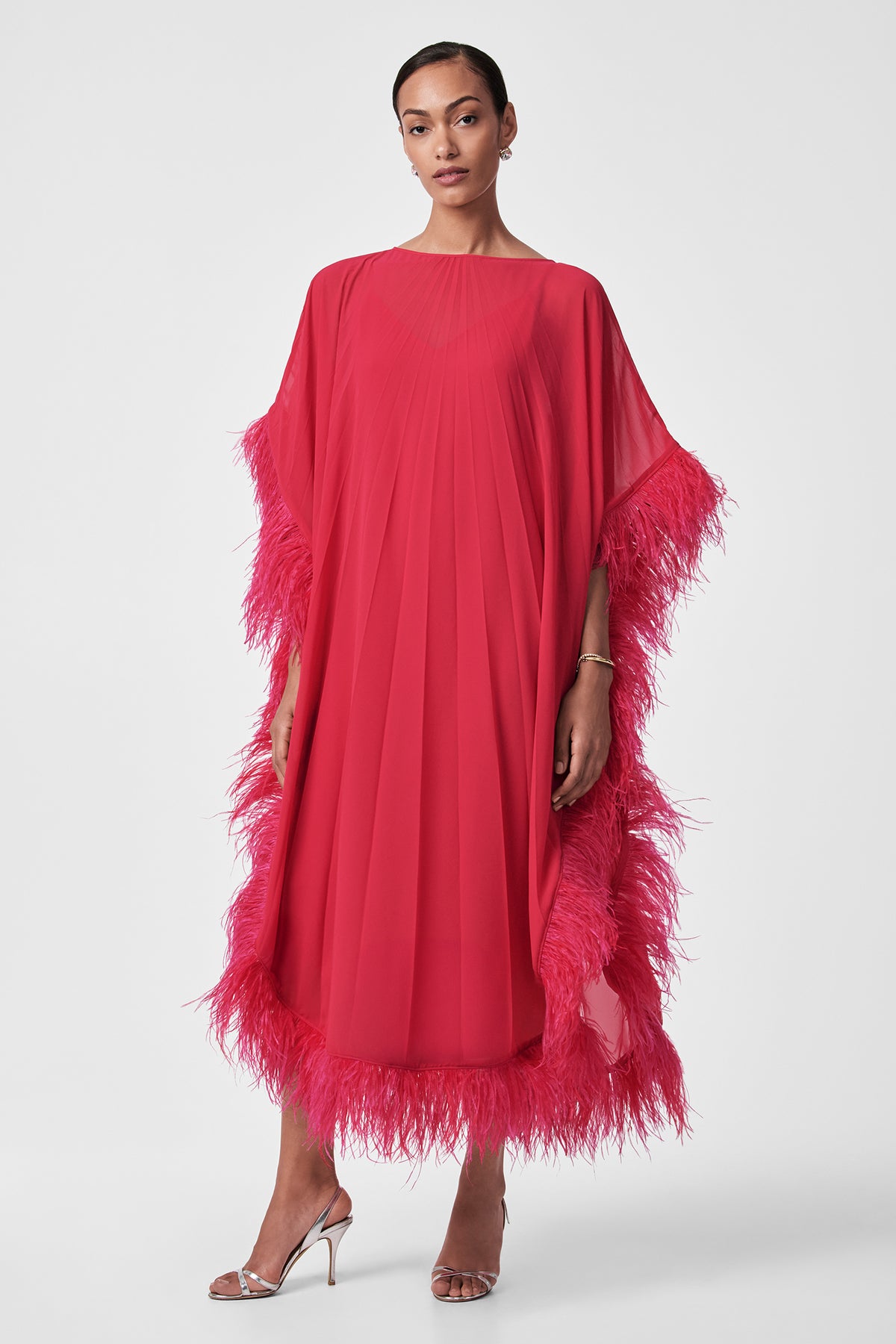 A woman poses against a white background in the Pleated Neena Feather-Trimmed Caftan Dress, a bright pink, loose-fitting style with pleats and feathered trim on the sleeves, hem, and sides. She pairs it with silver high-heeled sandals and wears her hair pulled back.