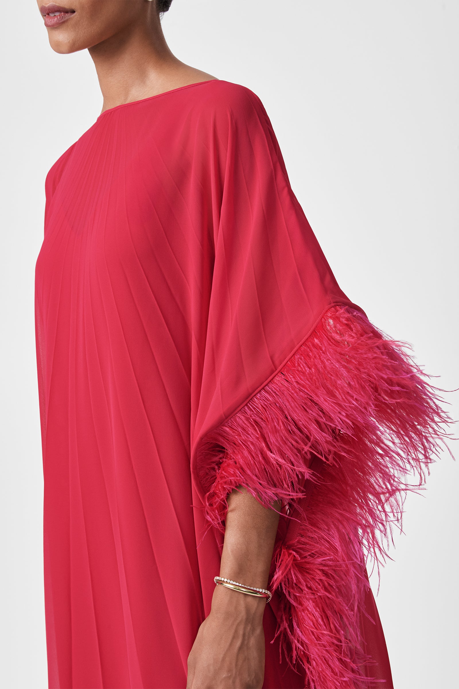 A woman in the Pleated Neena Feather-Trimmed Caftan Dress, featuring a bright red hue, pleats, and feather-trimmed sleeves. She has short hair, wears a thin gold bracelet, and stands against a plain white background.