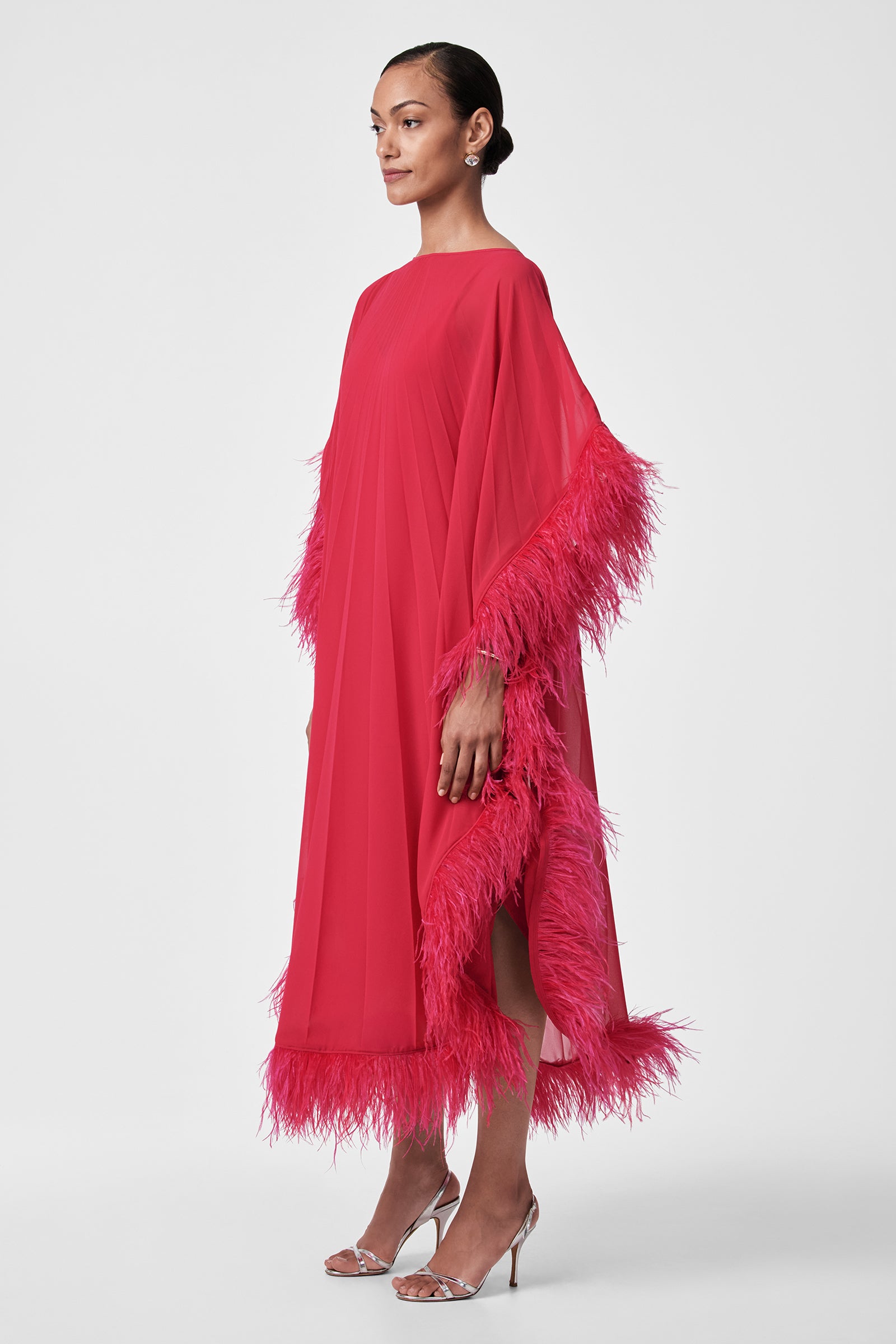 A person in profile models the Pleated Neena Feather-Trimmed Caftan Dress—loose, bright pink with feather trim on sleeves and hem—paired with silver high-heeled sandals against a plain white background.