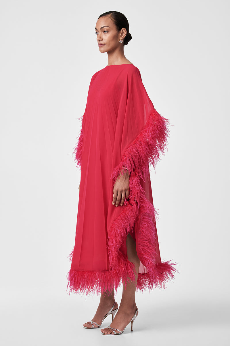 A person in profile models the Pleated Neena Feather-Trimmed Caftan Dressâ€”loose, bright pink with feather trim on sleeves and hemâ€”paired with silver high-heeled sandals against a plain white background.