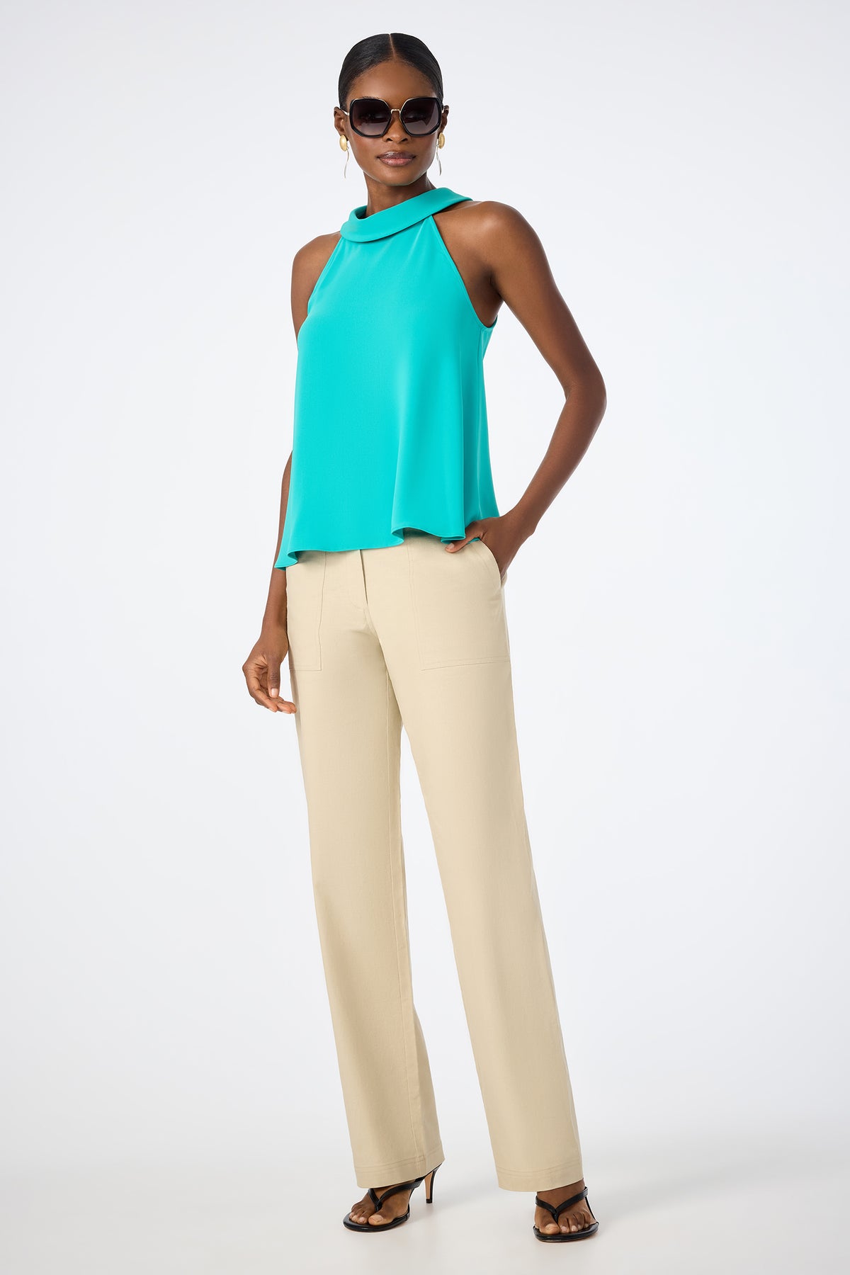 A woman stands confidently against a white background wearing the Eze Halter Top in teal, beige wide-leg trousers, black heeled sandals, large hoop earrings, and dark sunglasses.