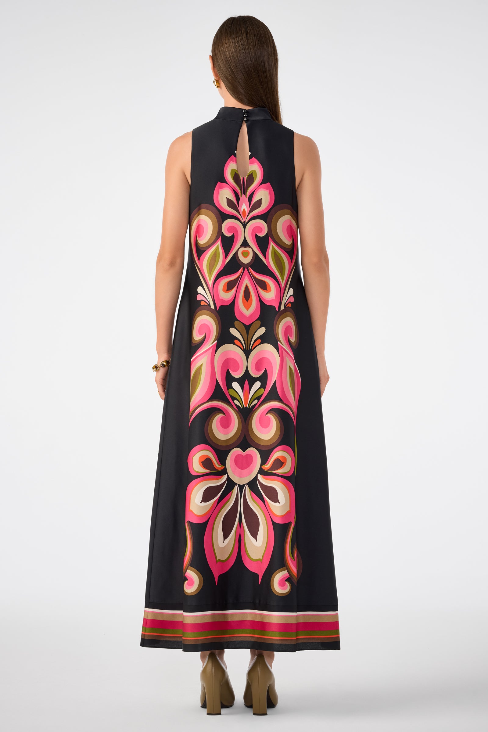 A woman with straight brown hair faces away from the camera, wearing the Loveage Sleeveless Maxi Dress—a black maxi dress featuring a striking pink, red, and cream floral pattern down the back—paired with high-heeled shoes.