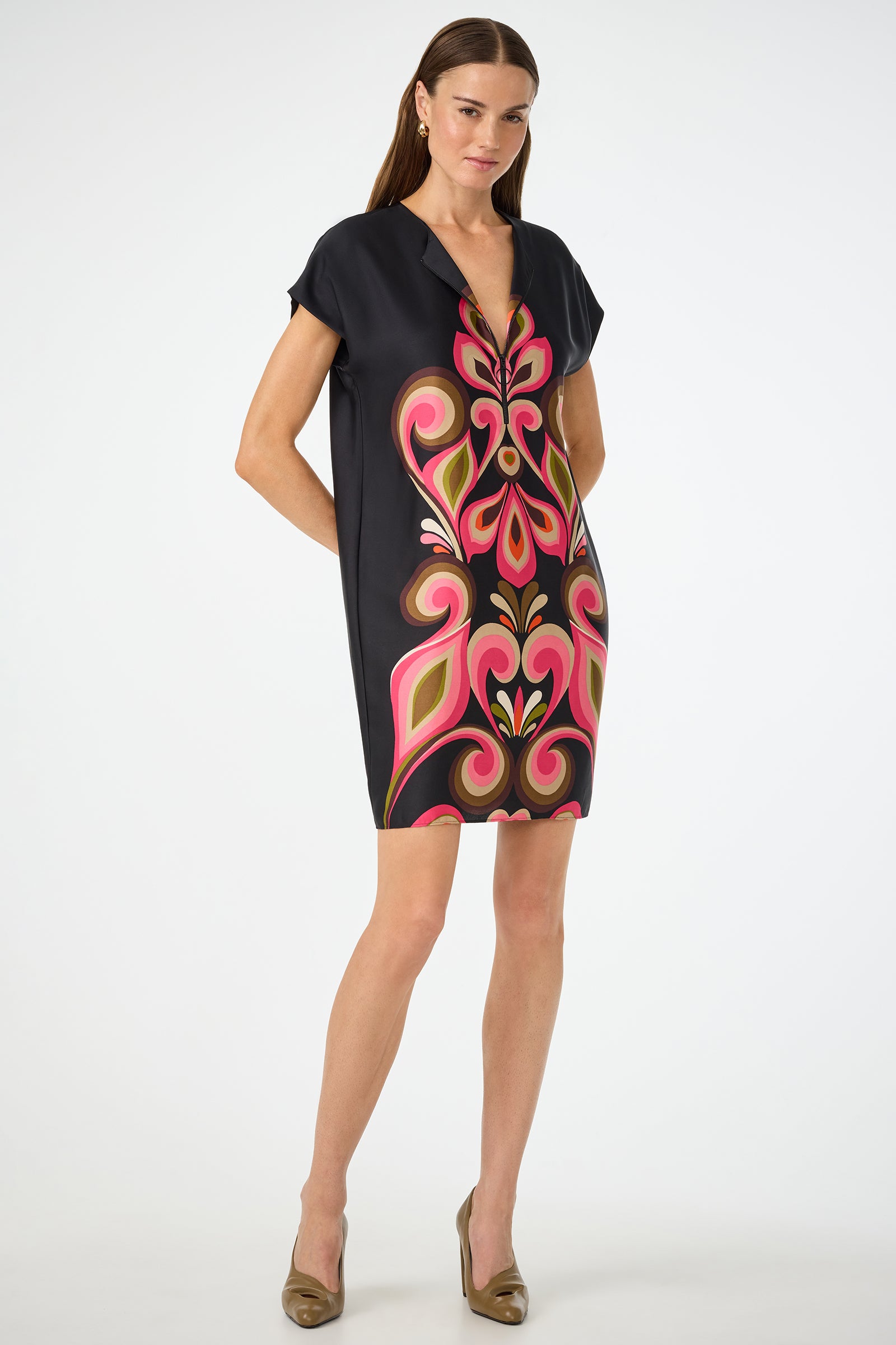 A woman with long brown hair and a neutral expression wears the Hasil 3 Short-Sleeve Shift Dress in Sanctuary, featuring a bold pink, red, and green abstract pattern on black, paired with taupe high heels.