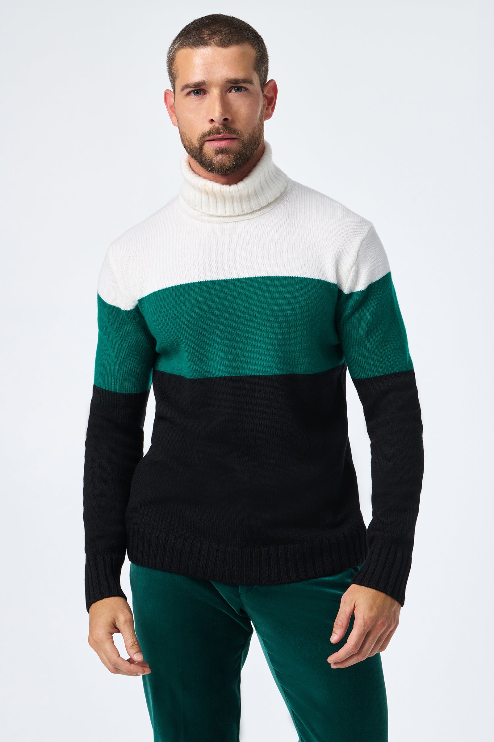 A man with short hair and a beard wears the Harrelson T-Neck, a colorblock sweater in white, green, and black, paired with green pants against a plain light background.