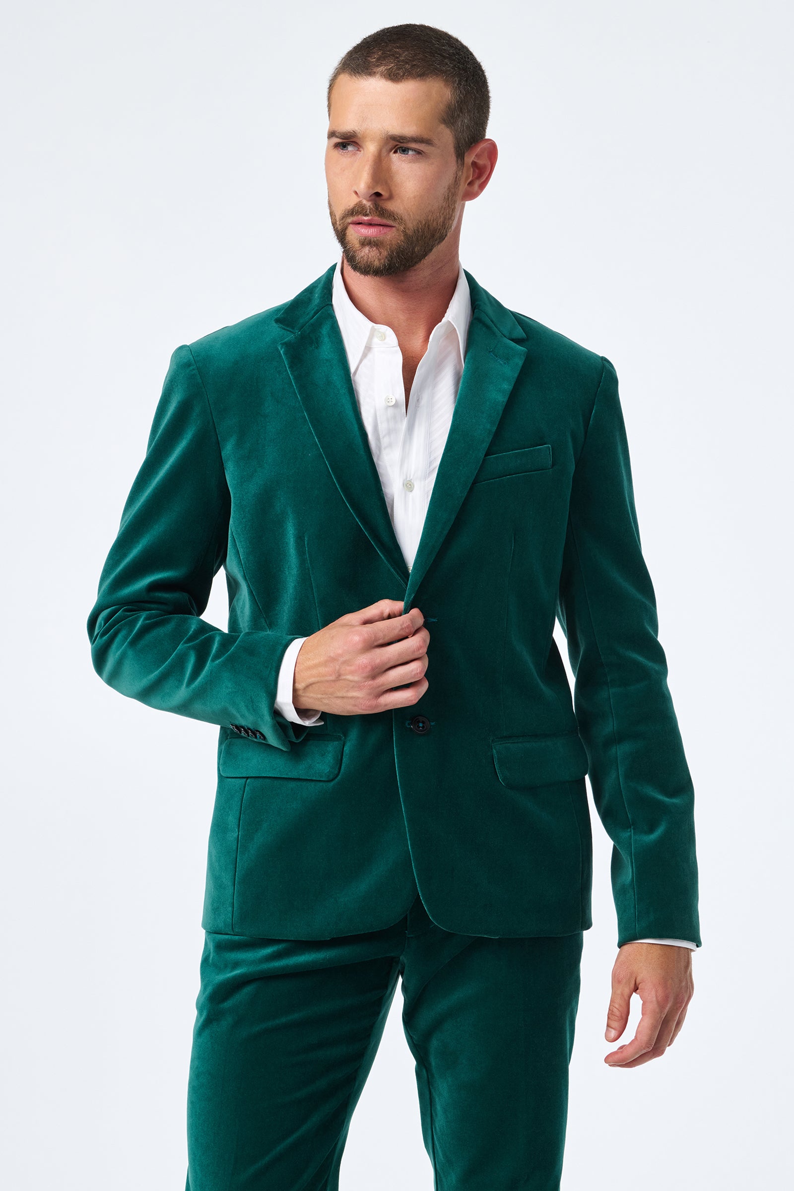 A man with short brown hair and a beard wears the Thurston Velvet Blazer in green over a white dress shirt, standing against a plain white background. He looks to the side while adjusting his jacket with one hand.
